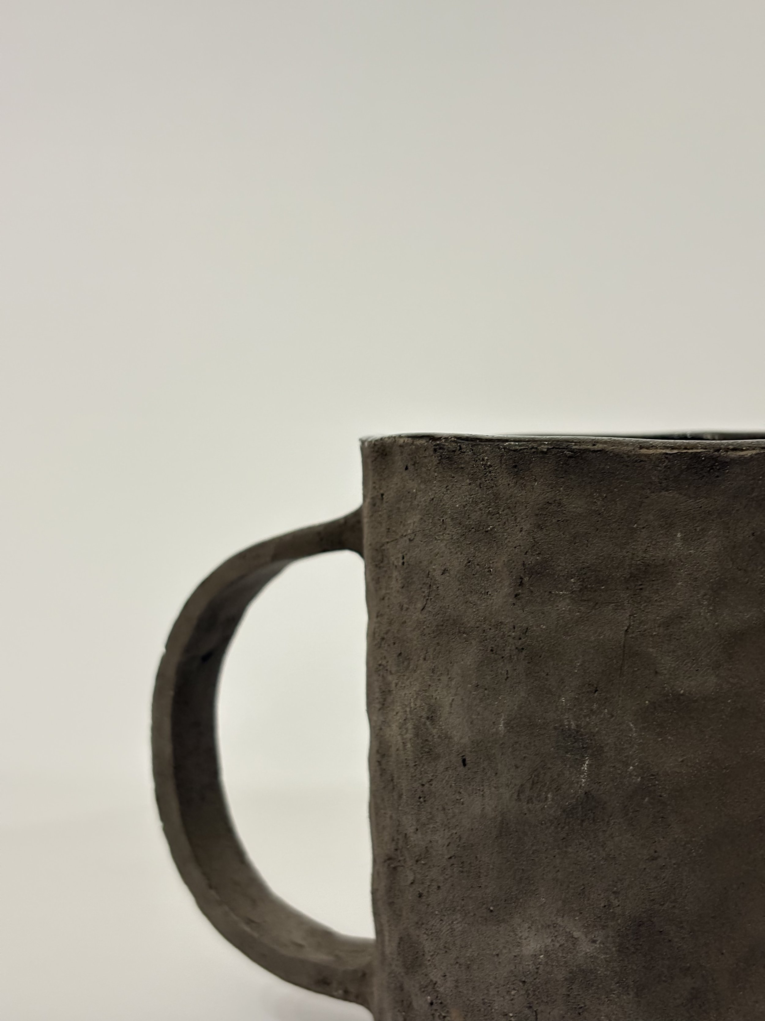 Una tazza di ceramica di colore grigio scuro con una superficie ruvida e un manico arrotondato, vista di lato su uno sfondo bianco.