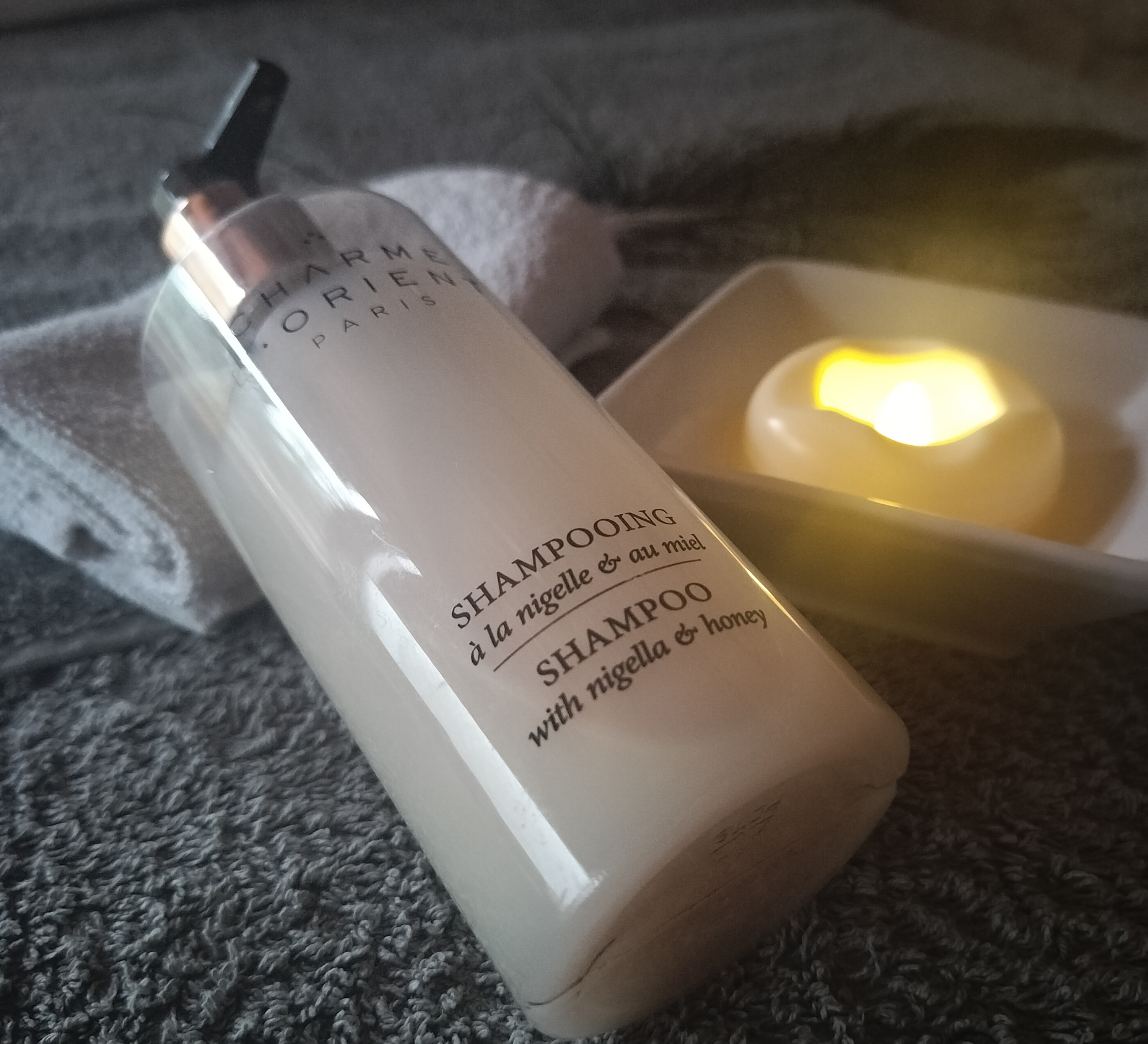 Bouteille de shampooing à la nigelle et au miel posée sur une serviette, avec une bougie allumée en arrière-plan dans un bol blanc.