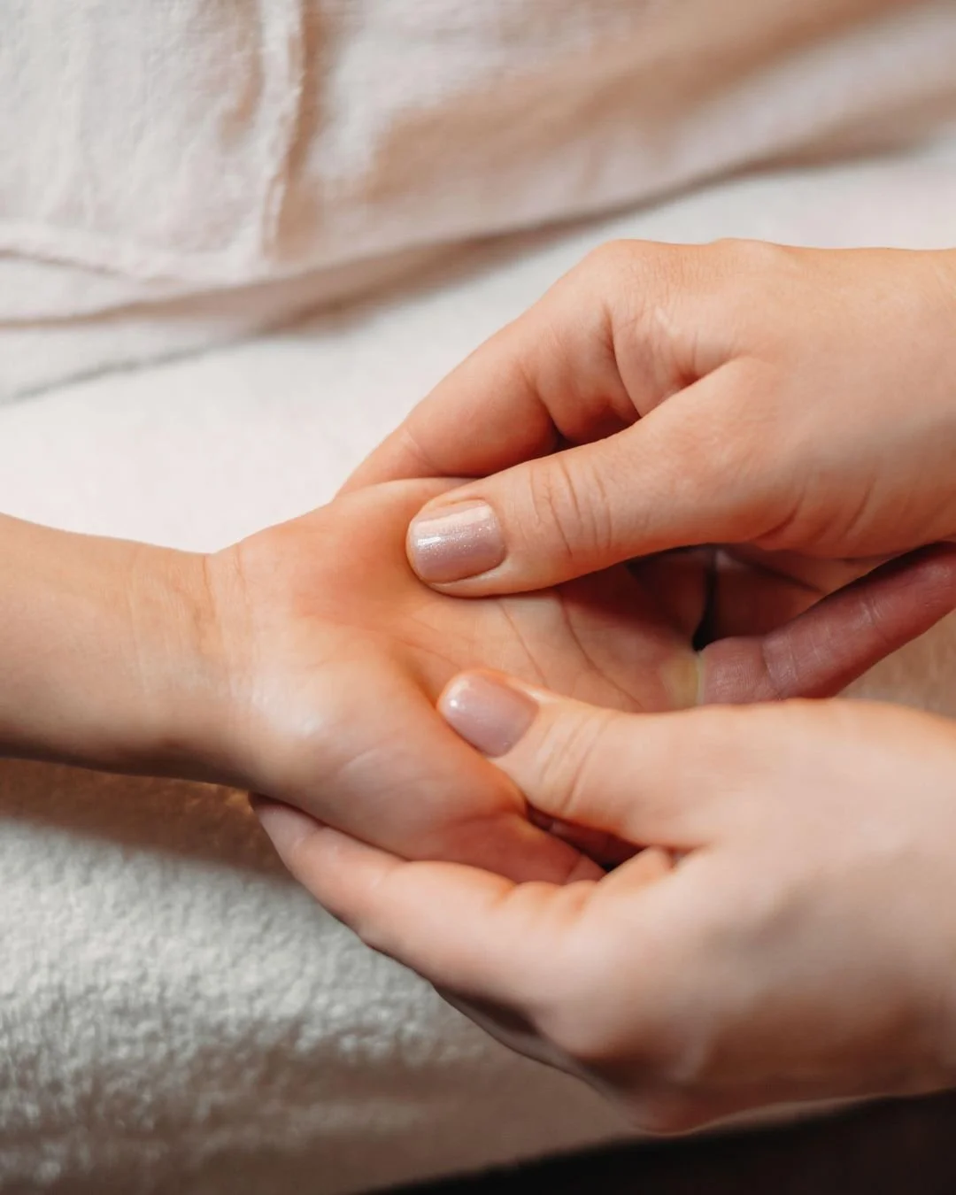 Deux mains tenant doucement une autre main, fond flou de tissu ou vêtement dans des tons rose ou beige.