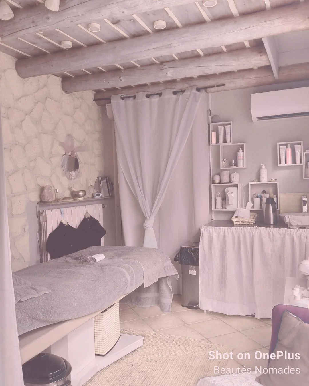 Salon de spa ou massage avec lit de massage, murs en pierre, étagères avec produits de beauté, rideau blanc, et mobilier confort.