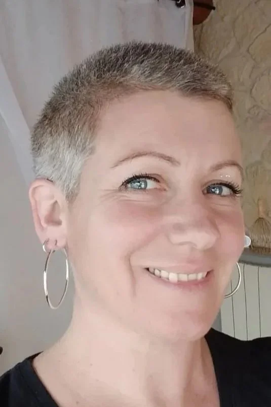 Une femme avec une coupe de cheveux très courte, souriante, portant de grandes boucles d'oreilles en anneau, en intérieur.
