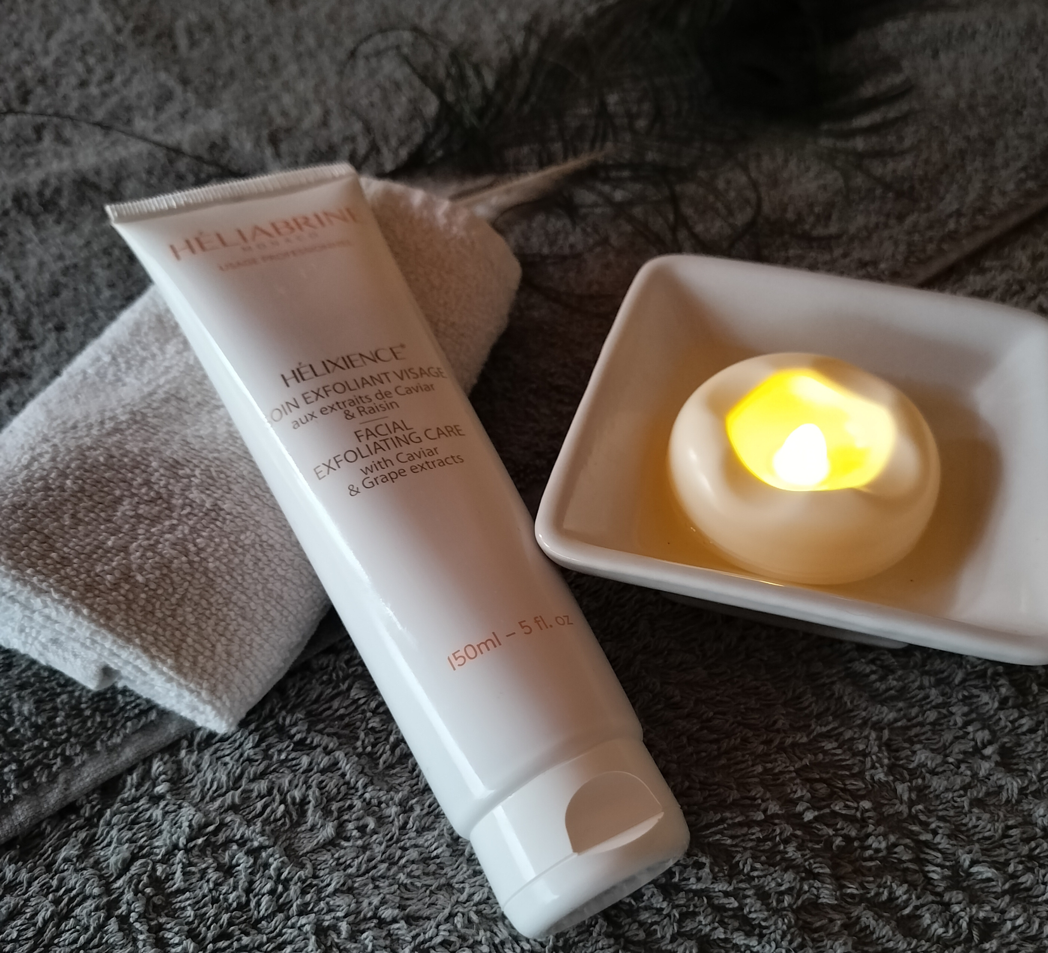 Tube de produit de soin facial, serviette blanche, bougie dans un plat blanc, sur une surface en tissu gris foncé.