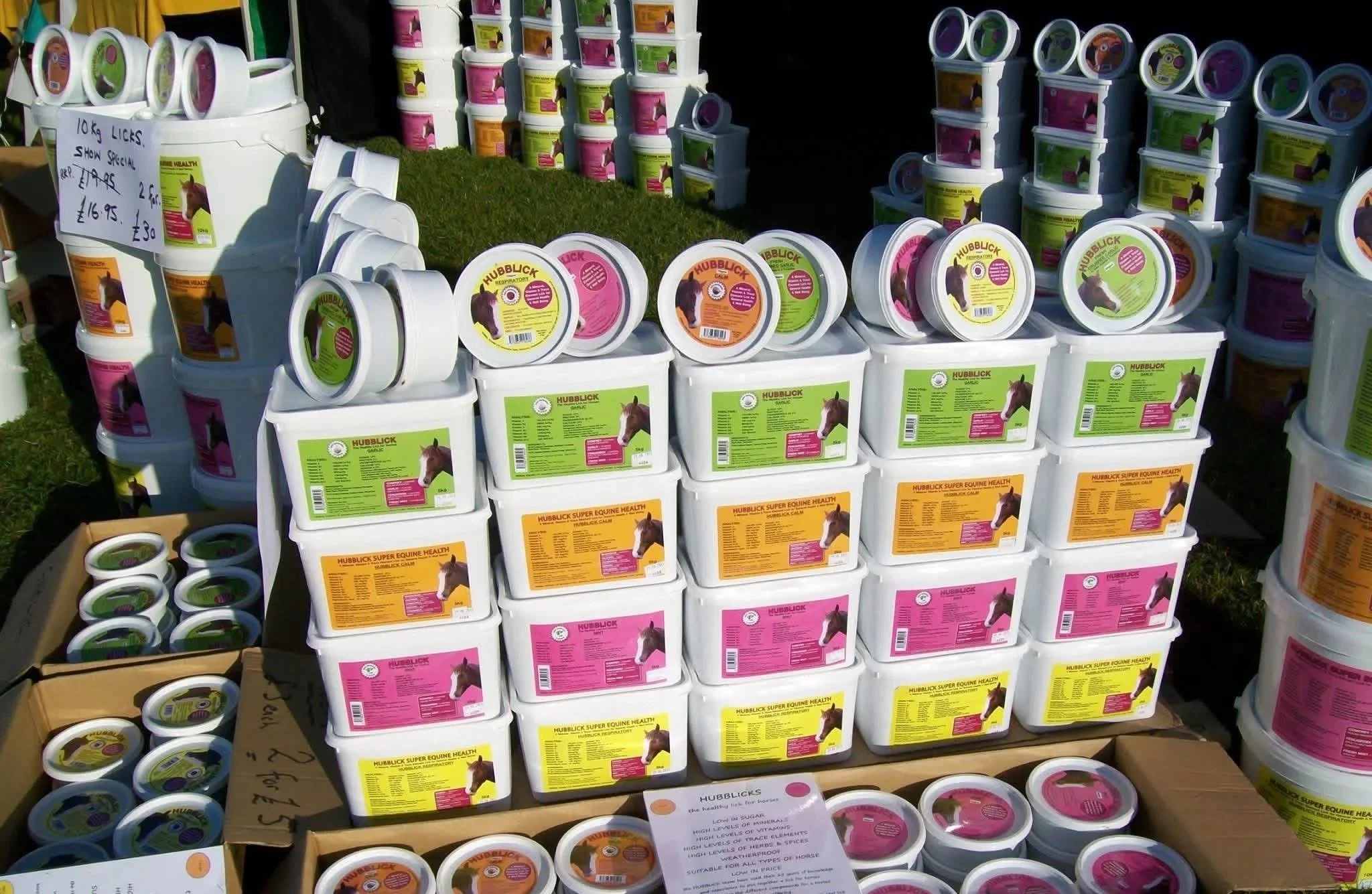 Assorted buckets of horse feed at an outdoor market, with various colorful labels and a handwritten sign.