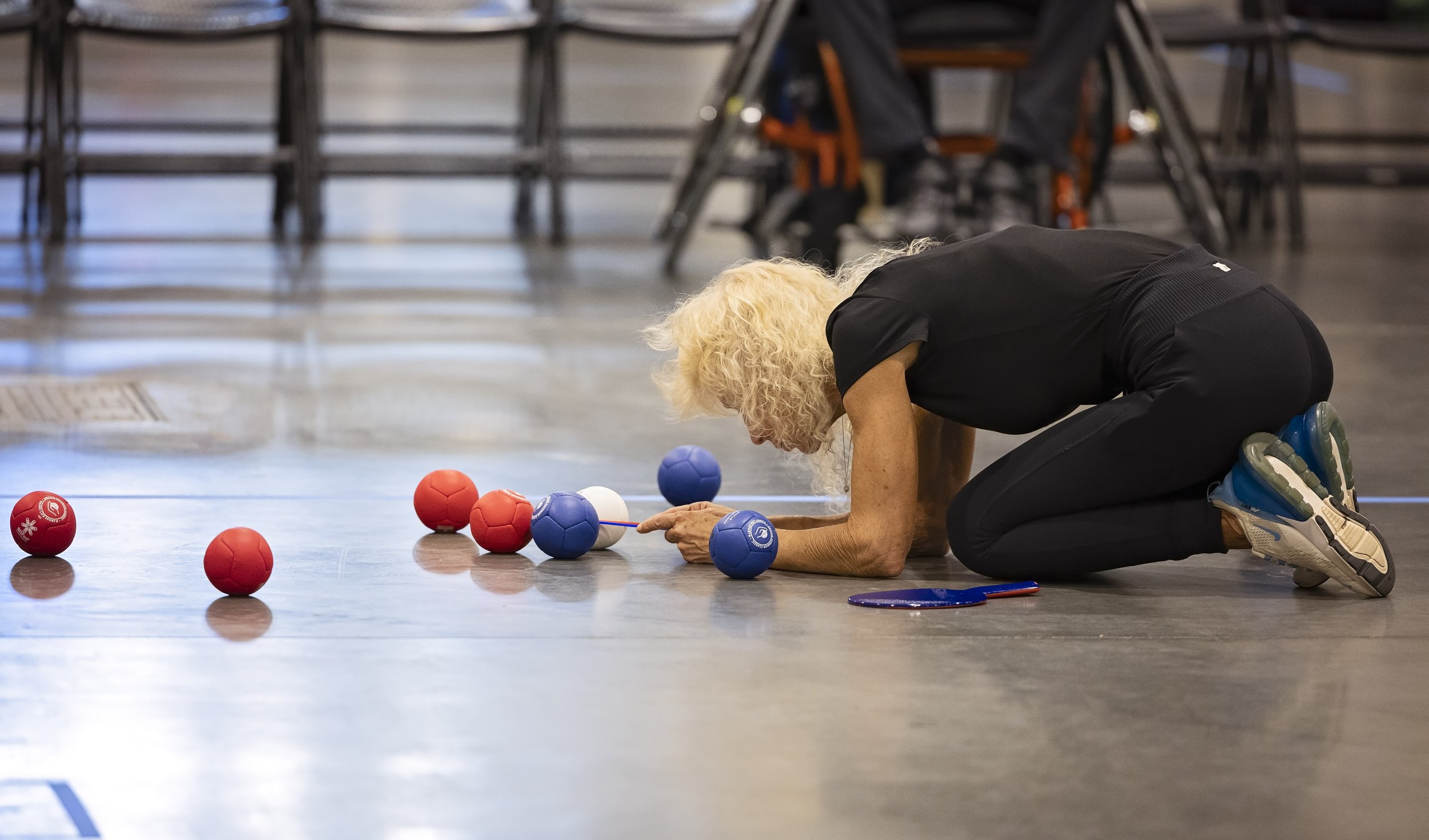 An older woman with curly blonde hair lying on the floor in a gymnasium, reaching towards colorful balls scattered on the floor, with a badminton racket nearby.