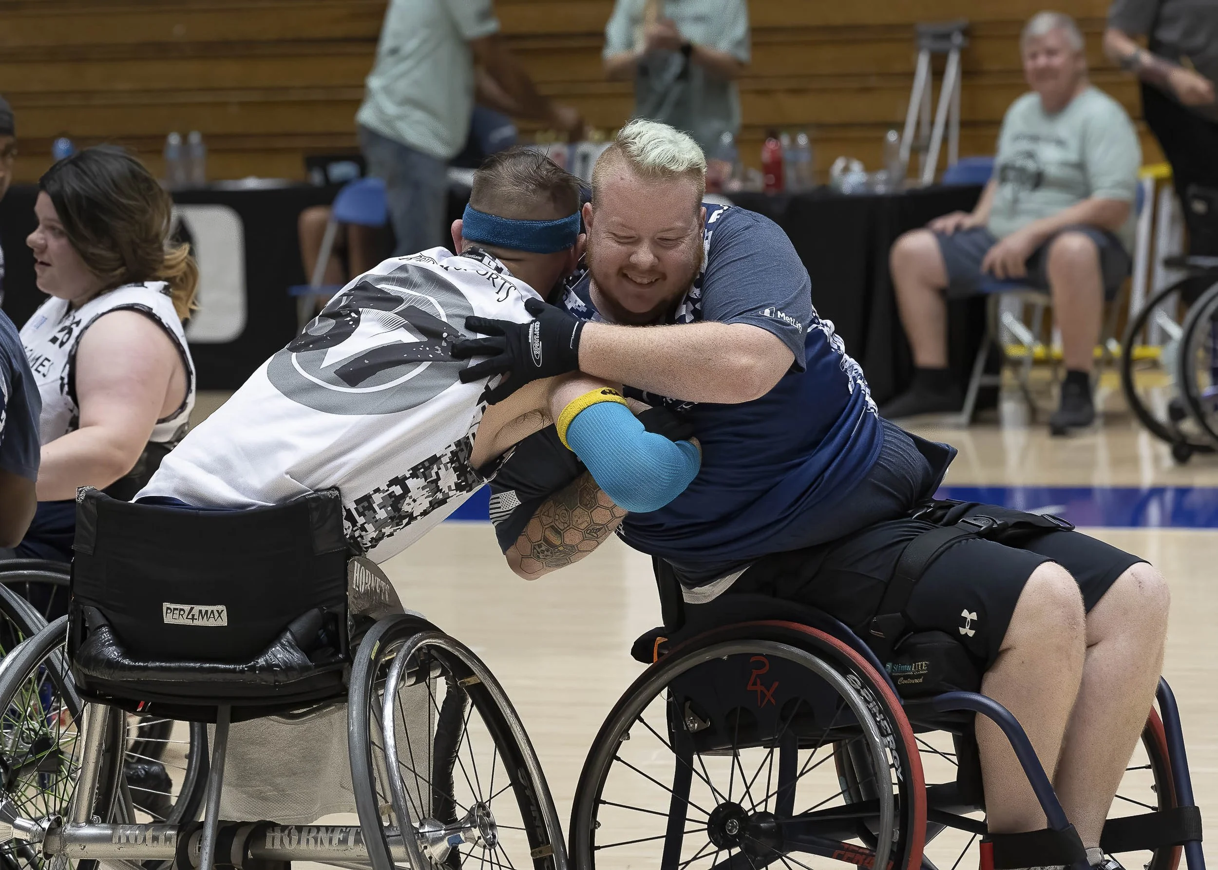 Two men in wheelchairs shaking hands and hugging in celebration on an indoor court, with other people in the background.