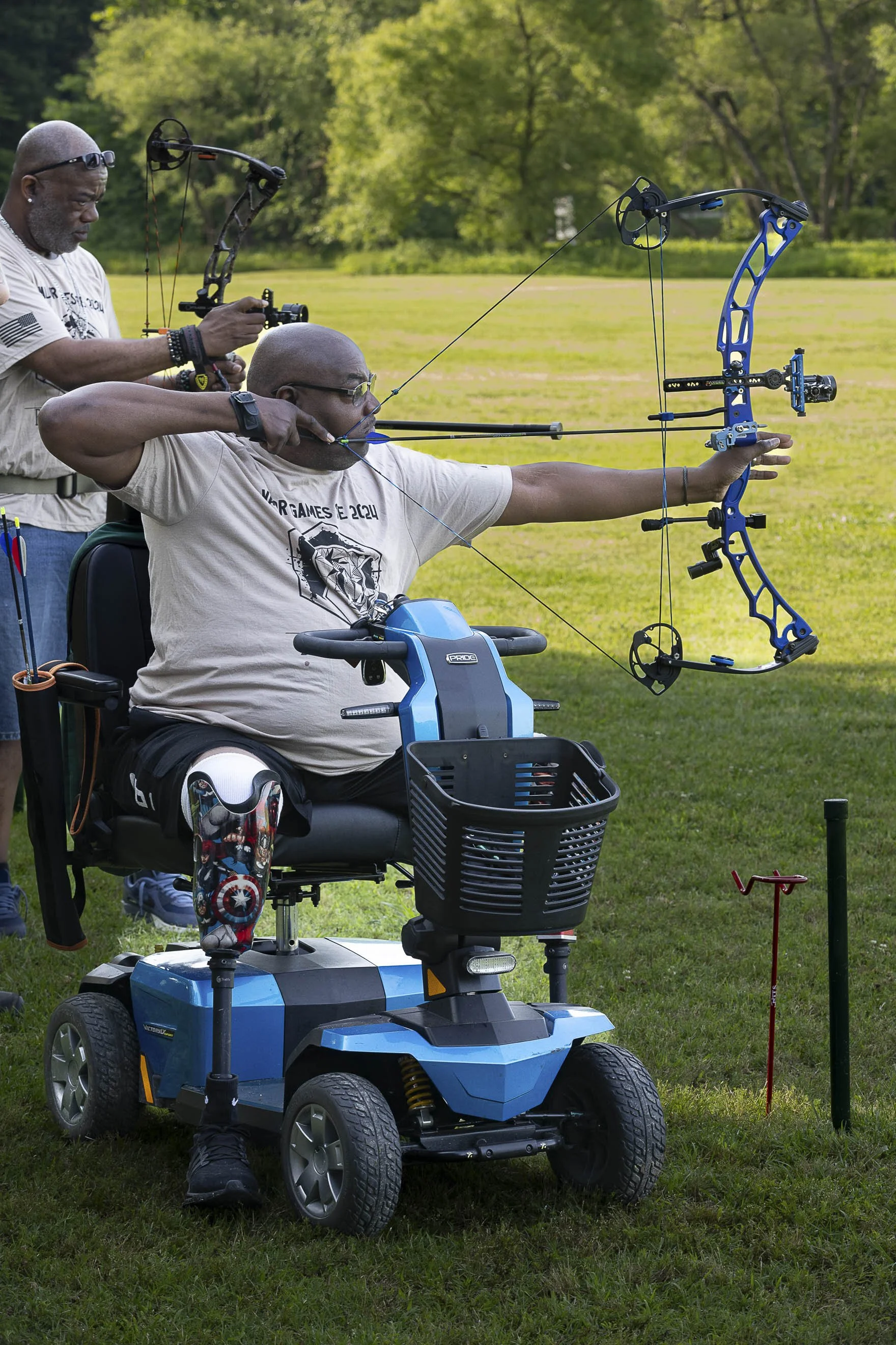 A man in a mobility scooter aiming a blue compound bow, assisted by another man standing behind him, on a grassy field with trees in the background.