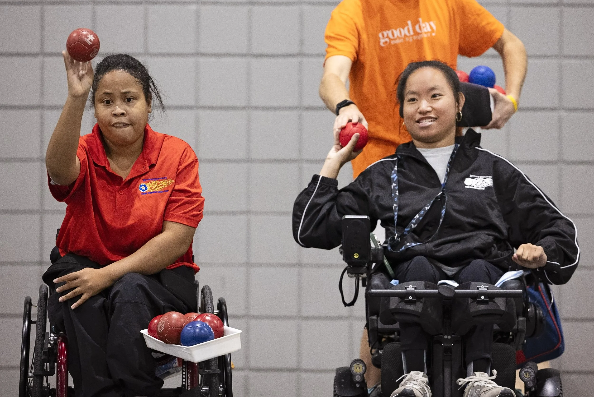 Two women in wheelchairs playing boccia with small balls, assisted by a man standing behind them.