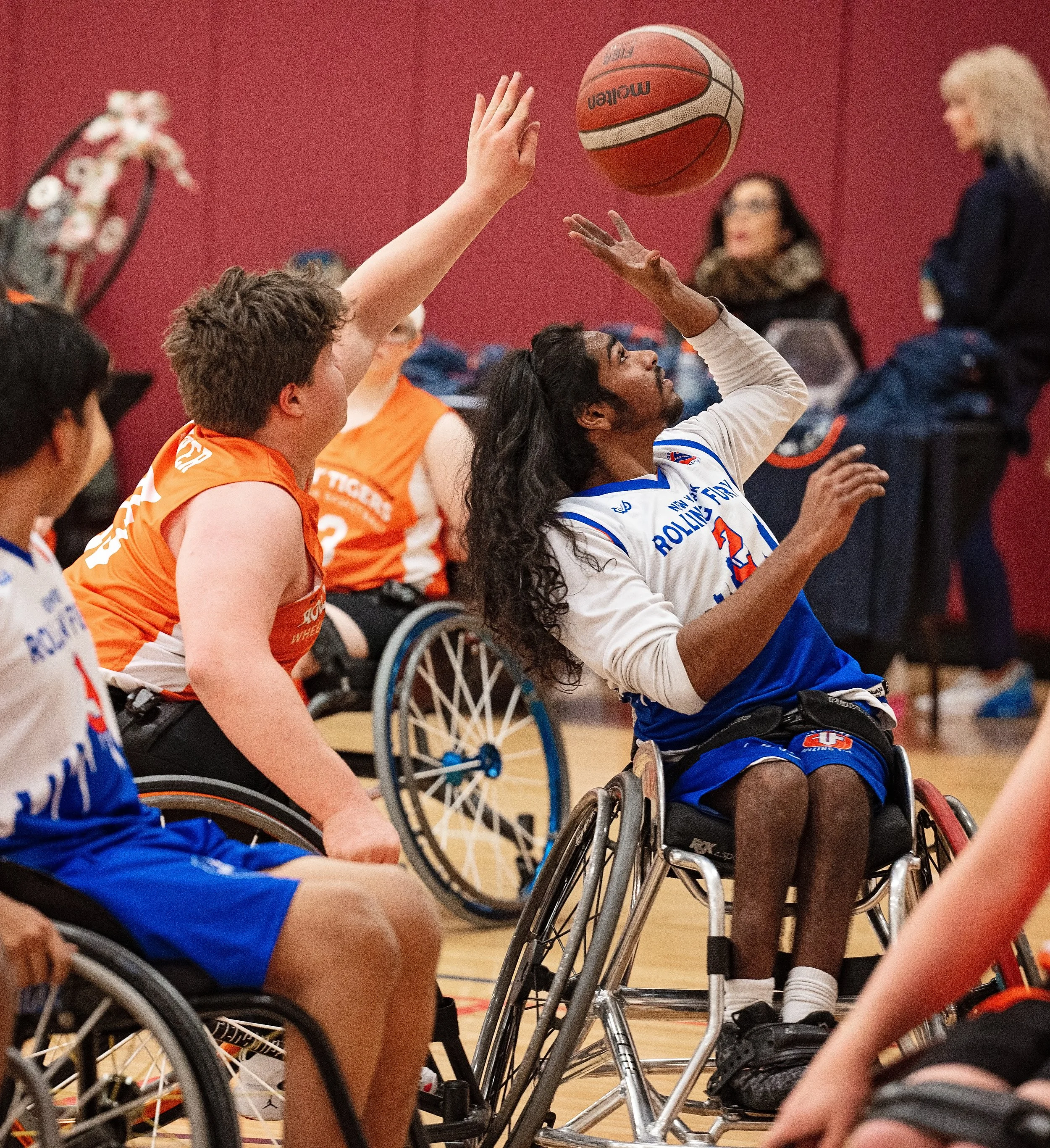 Wheelchair basketball players in action during a game, with players wearing jerseys in white, blue, and orange.