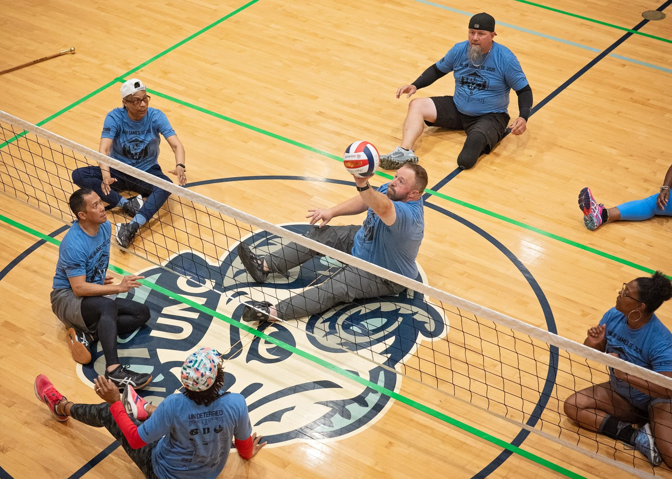People playing sitting volleyball inside a gymnasium on a wooden floor, with a logo on the court, some participants sitting and others reaching to hit a volleyball over the net.