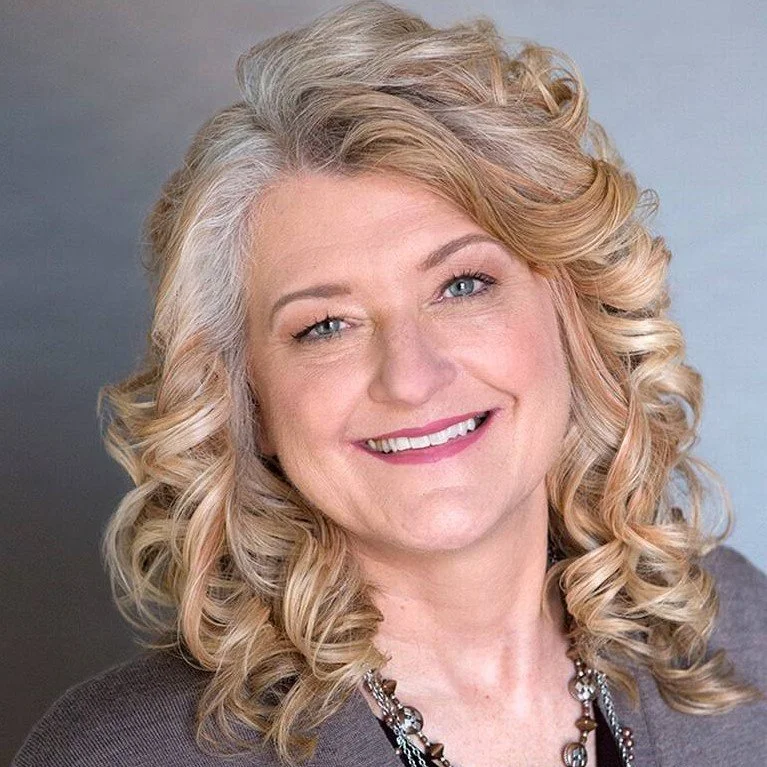 A woman with blonde, curly hair smiling at the camera, wearing a dark top and a necklace.