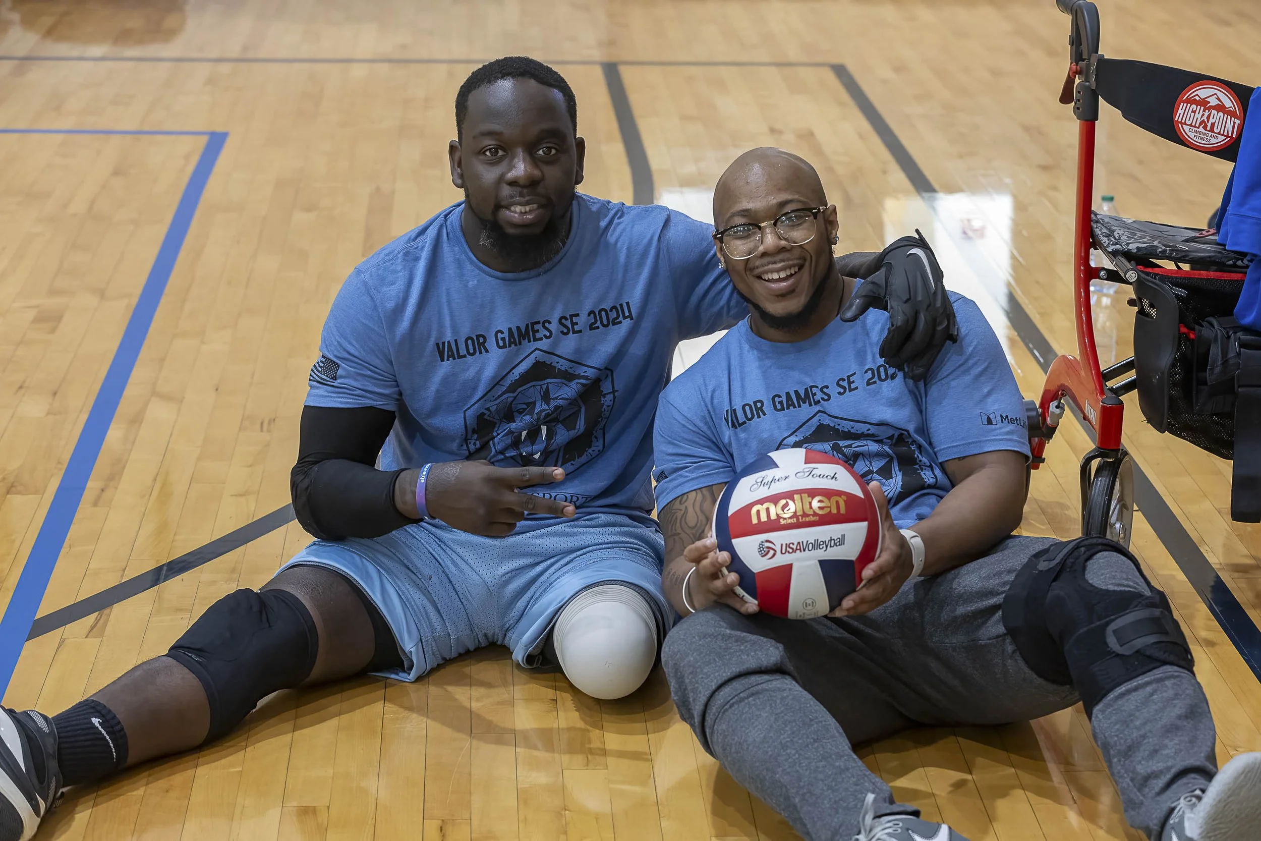 Two men sitting on the indoor volleyball court floor, wearing matching blue Valor Games SE 2024 t-shirts, one with a leg injury resting on a foam roller, holding a volleyball, smiling at the camera, with sports equipment nearby.