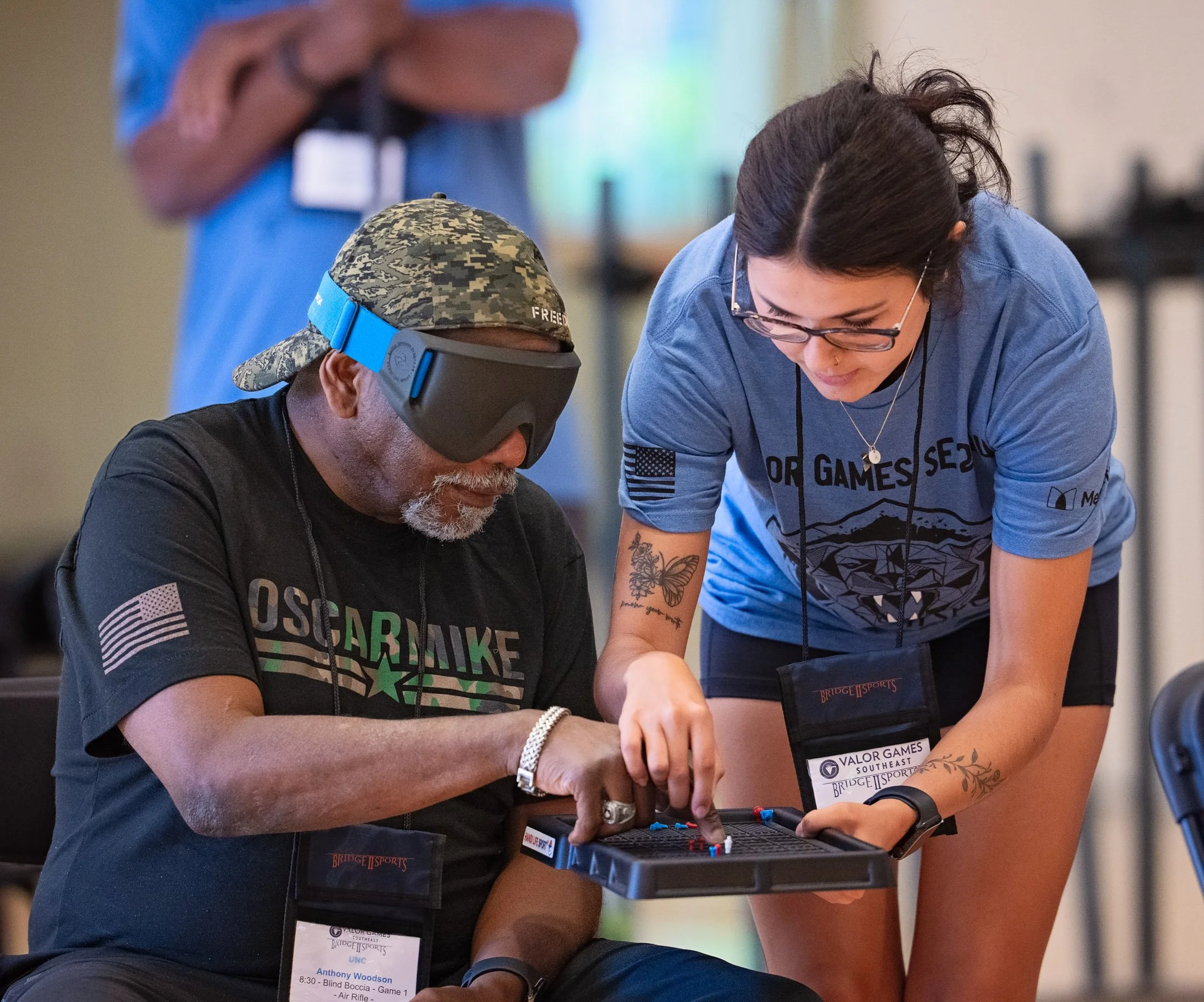 A woman in a blue T-shirt and glasses assists a man in a black T-shirt with a virtual reality headset, as they play a tabletop game.