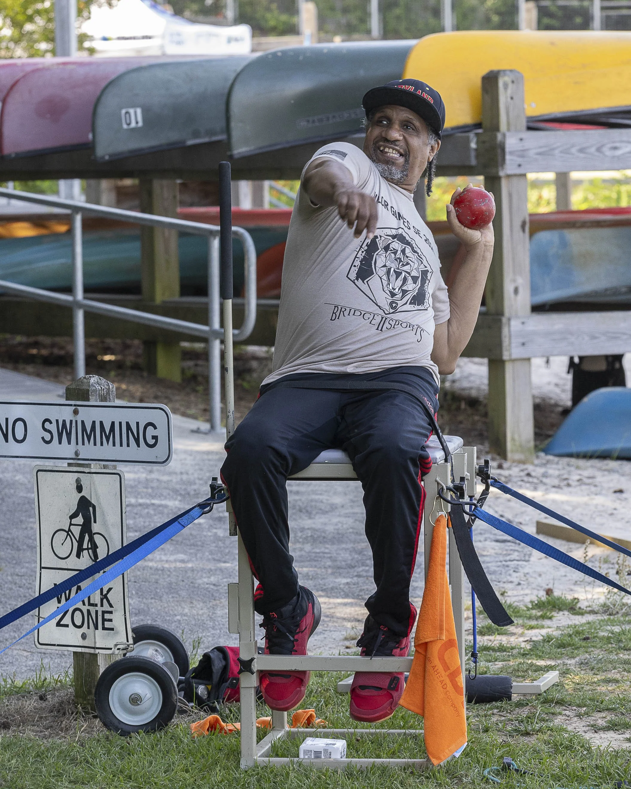A man sitting in a shot-put chair outdoors near canoes stacked on racks, competing at Valor Games Southeast
