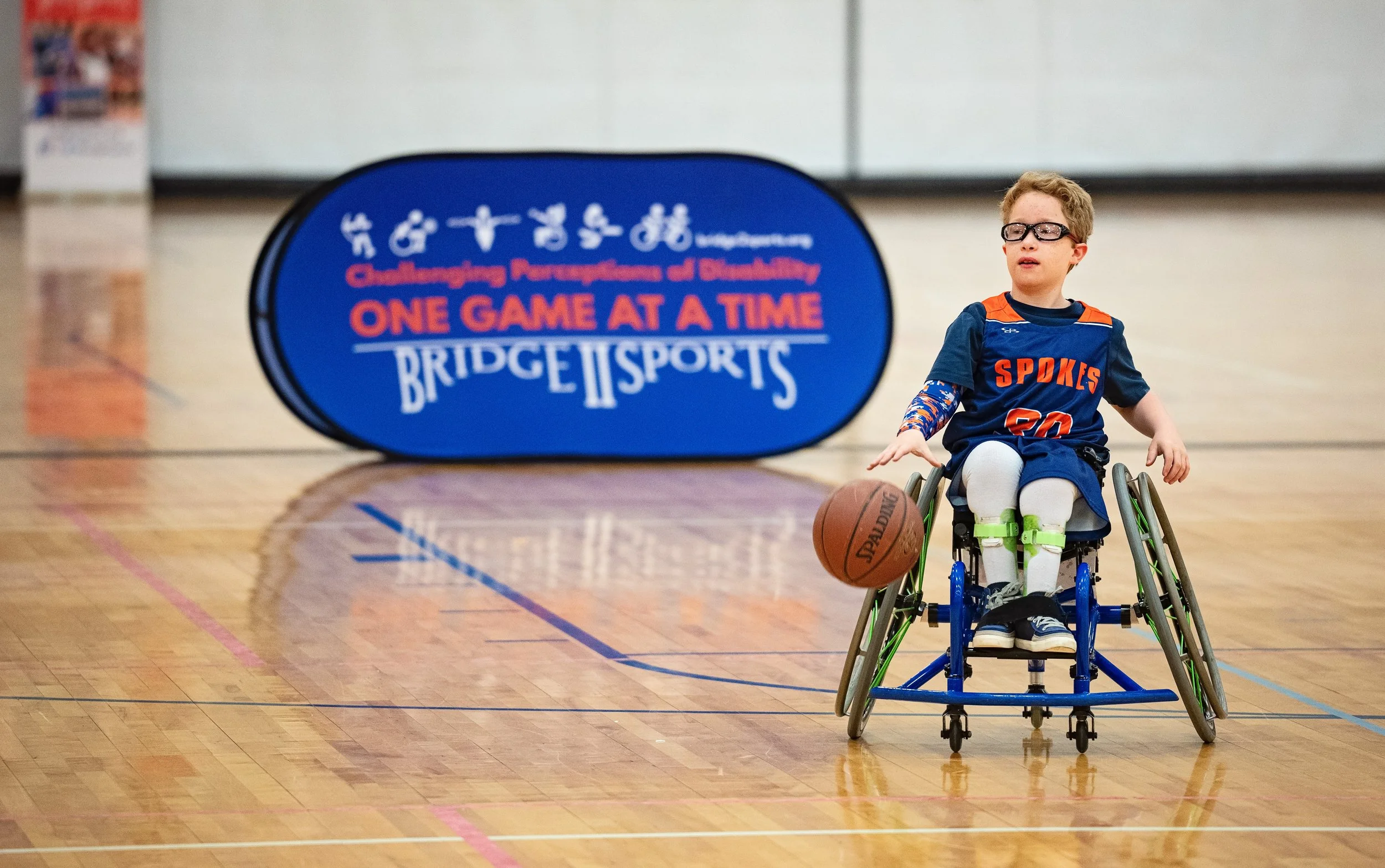 A young boy in a wheelchair on a basketball court holding a basketball with a sign in the background that reads 'Challenging Perceptions of Disability' and 'One Game at a Time' related to bridge sports.