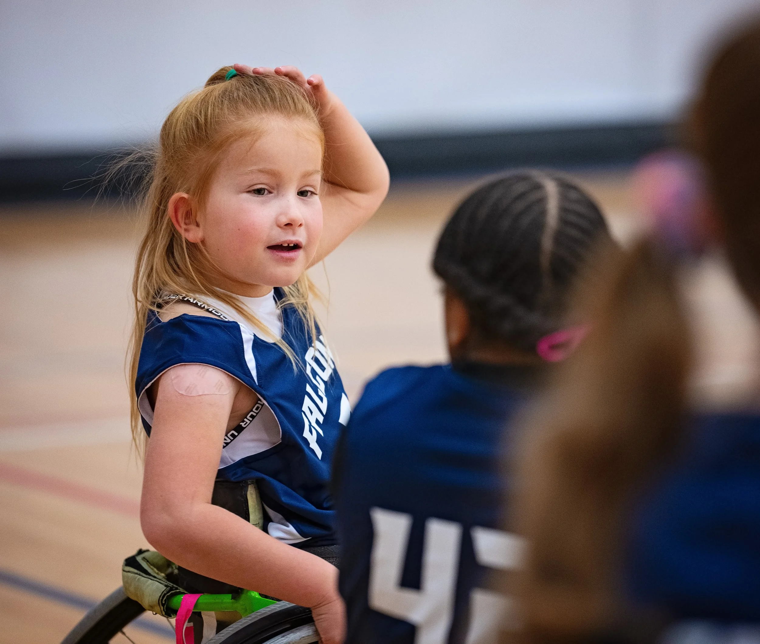 A young girl with red hair in a wheelchair, wearing a sports jersey, talking to a group of people in a gymnasium.