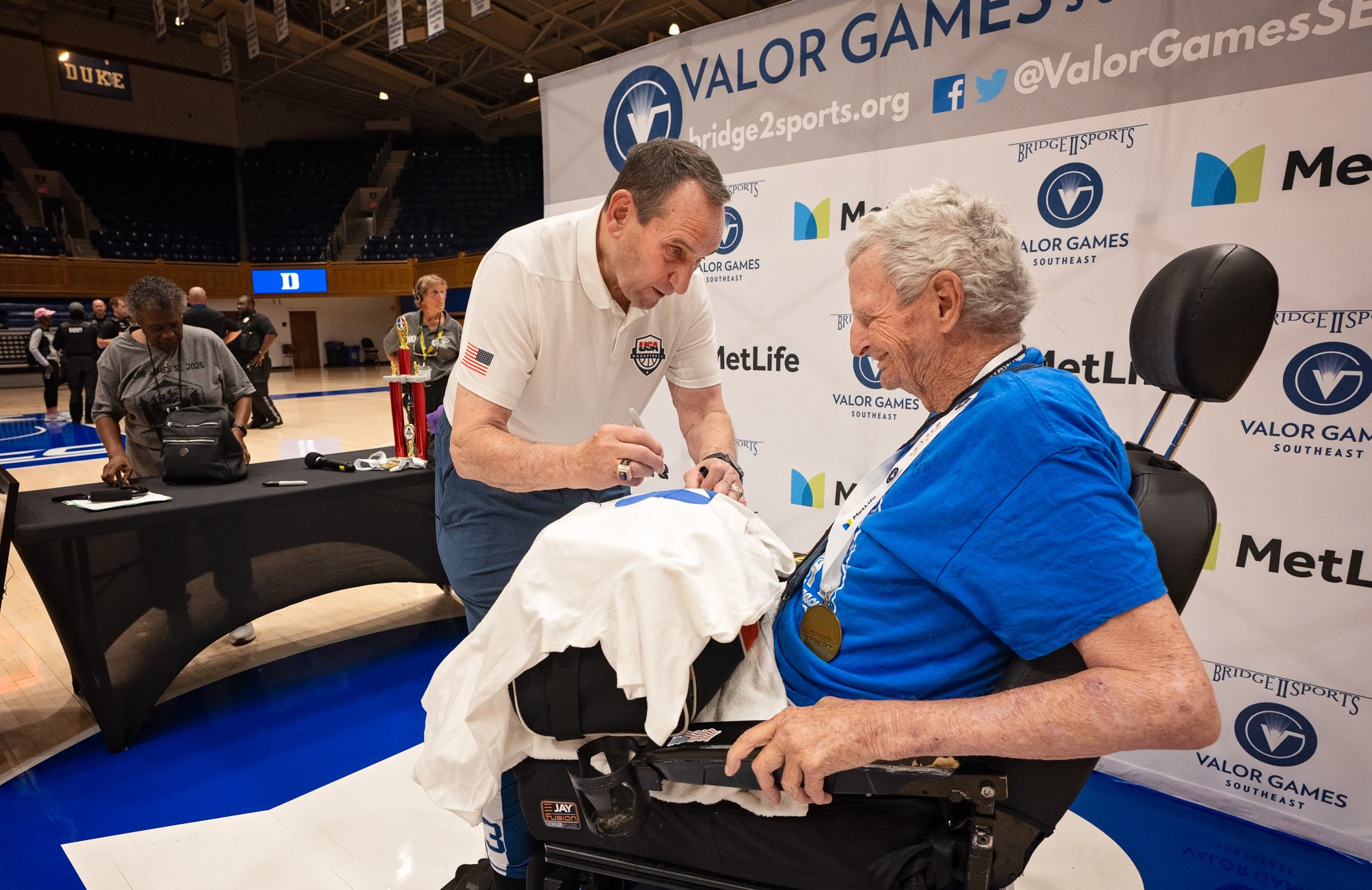 An elderly man with gray hair and a blue shirt is sitting in a wheelchair, smiling and receiving a signature from Mike Krzyzewski at the Valor Games.