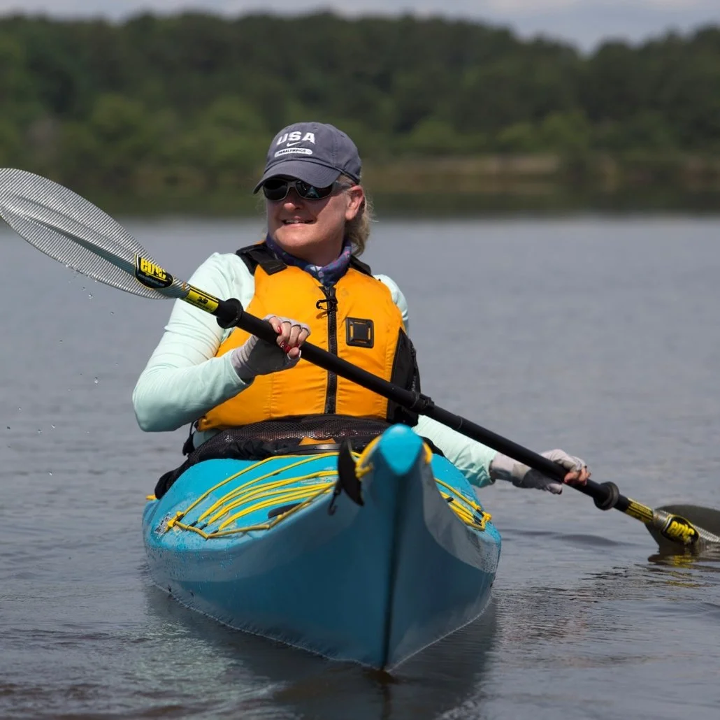 A woman kayaking on a lake, wearing sunglasses, a cap with 'USA' on it, a yellow life jacket, and a light-colored long-sleeve shirt, holding a paddle.