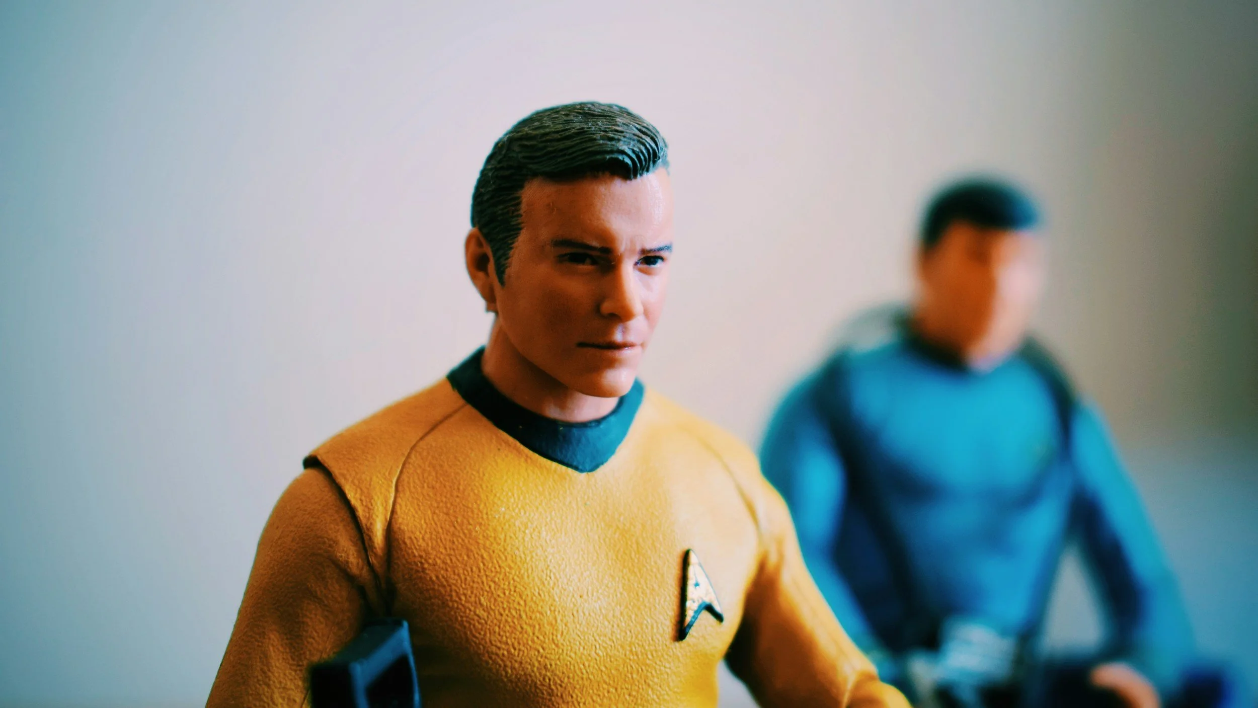 Close-up of a male action figure in a yellow Star Trek uniform with a Starfleet insignia, standing in front of a blurred background with another figure in a blue uniform.