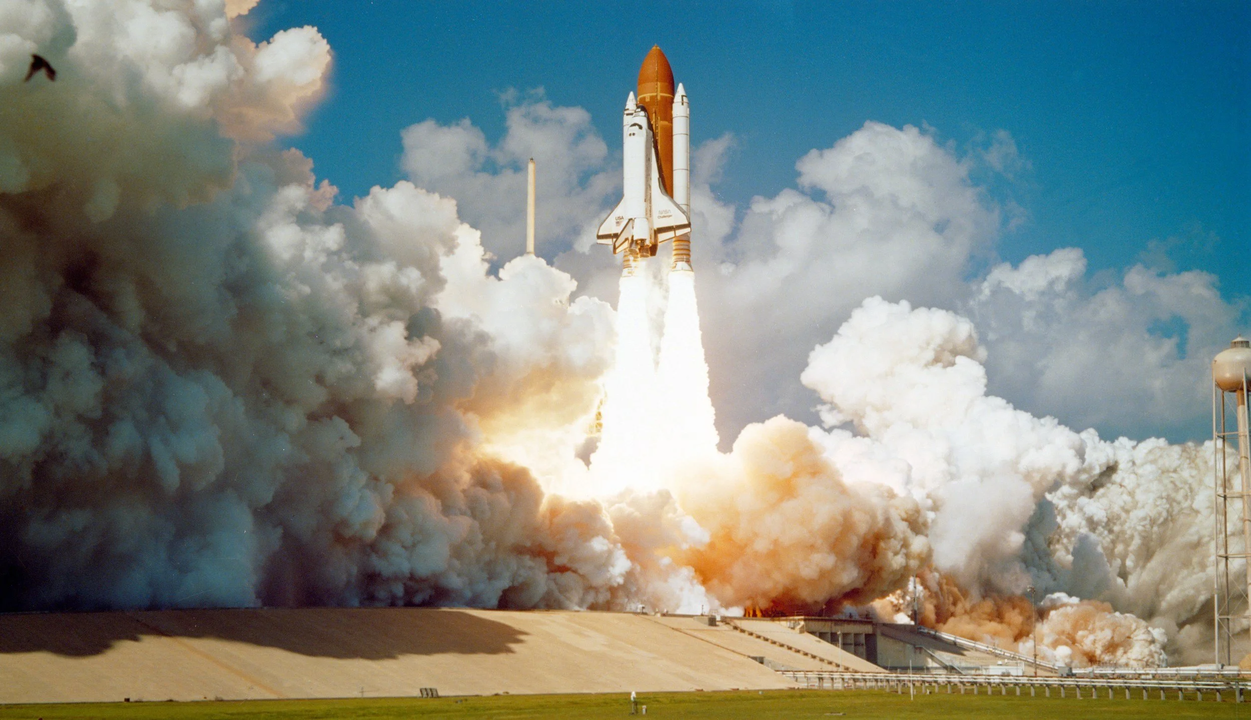 NASA space shuttle launches with orange external tank and white solid rocket boosters, creating a large cloud of smoke and flames.