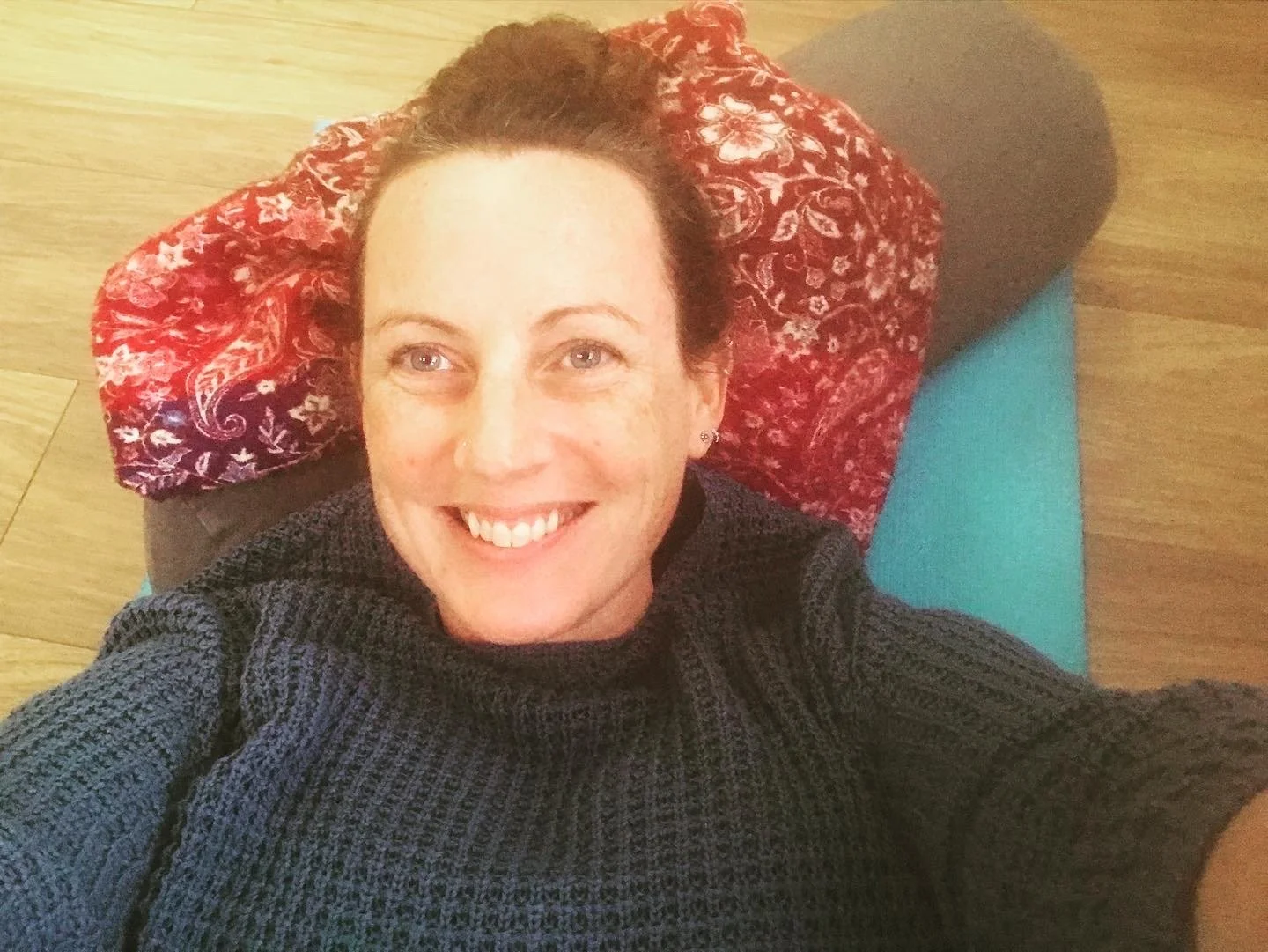 A woman with short brown hair smiling, lying on a red floral pillow and a teal yoga mat on a wooden floor, wearing a dark gray sweater.