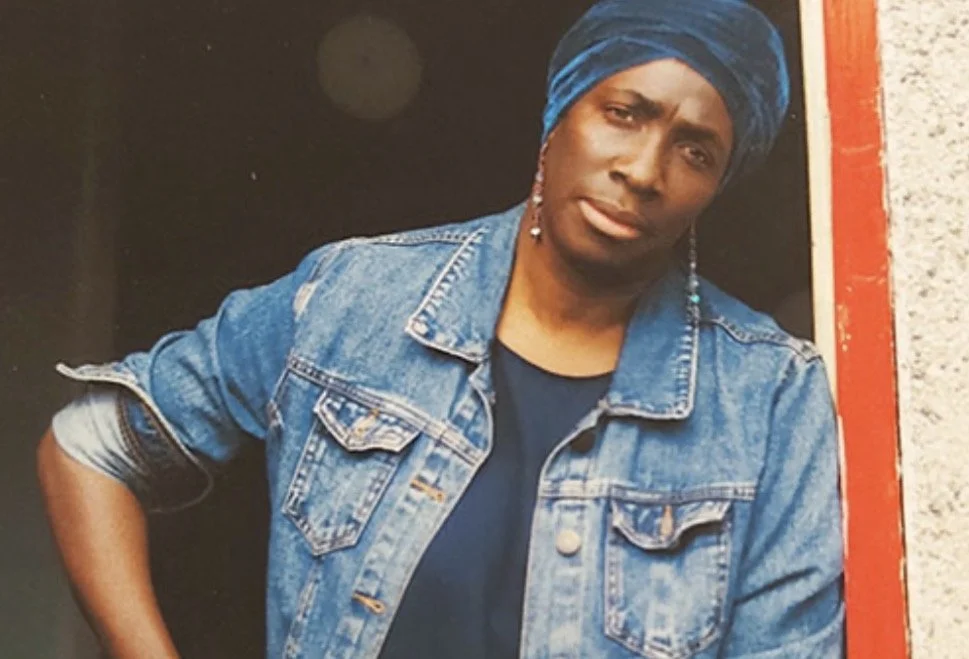 A woman with a dark blue headwrap, denim jacket, and earrings standing against a dark background.