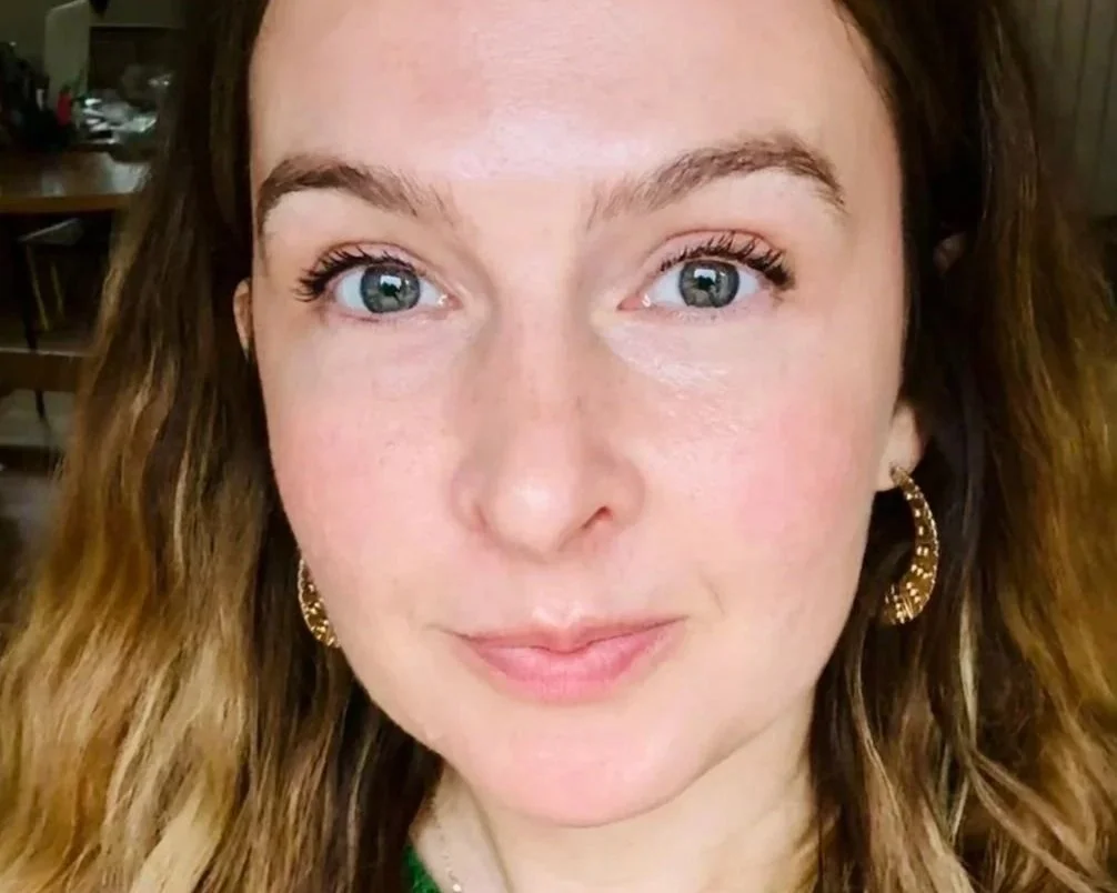 Close-up of a woman with blue eyes, light skin, brown wavy hair, wearing gold hoop earrings, in a room with wooden furniture.