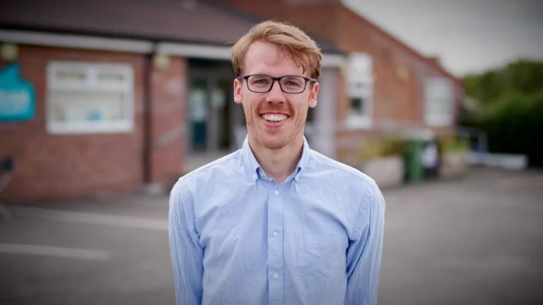 A young man with glasses and light brown hair smiling outdoors in front of a brick building.