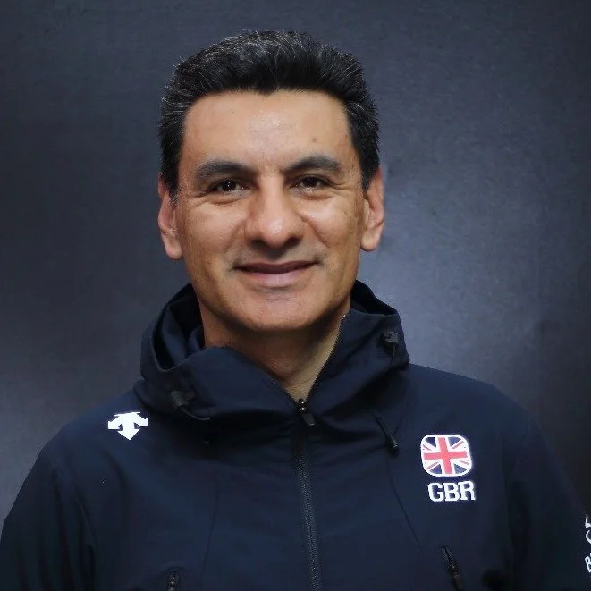 A man with dark hair smiling, wearing a navy blue jacket with the Union Jack flag and 'GBR' on the chest, standing against a dark backdrop.