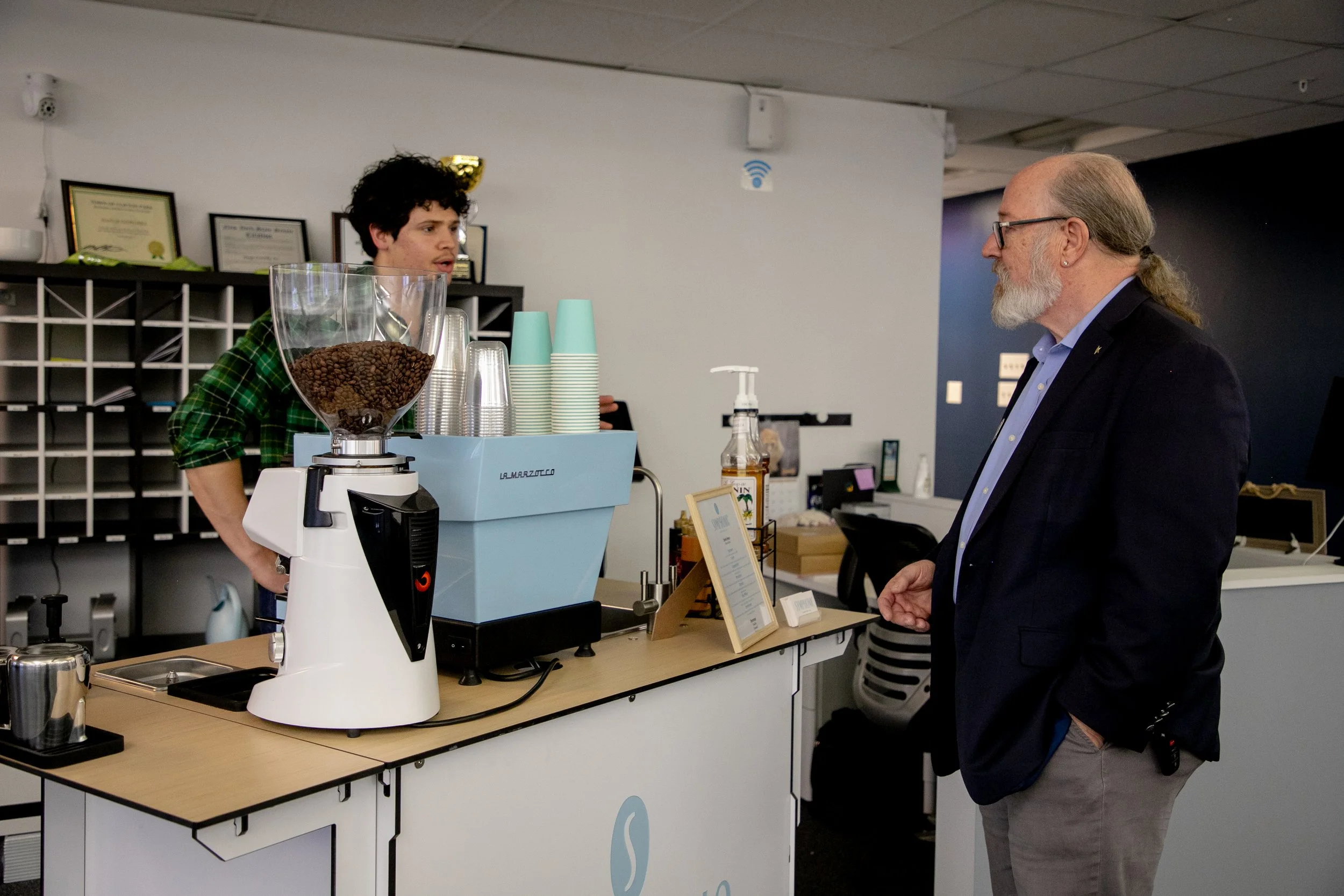 A man with gray hair and a beard speaking to a young male barista behind a coffee counter in a coffee shop. The barista is wearing a green plaid shirt and is standing next to a coffee grinder filled with coffee beans. The counter has a small sign, a 