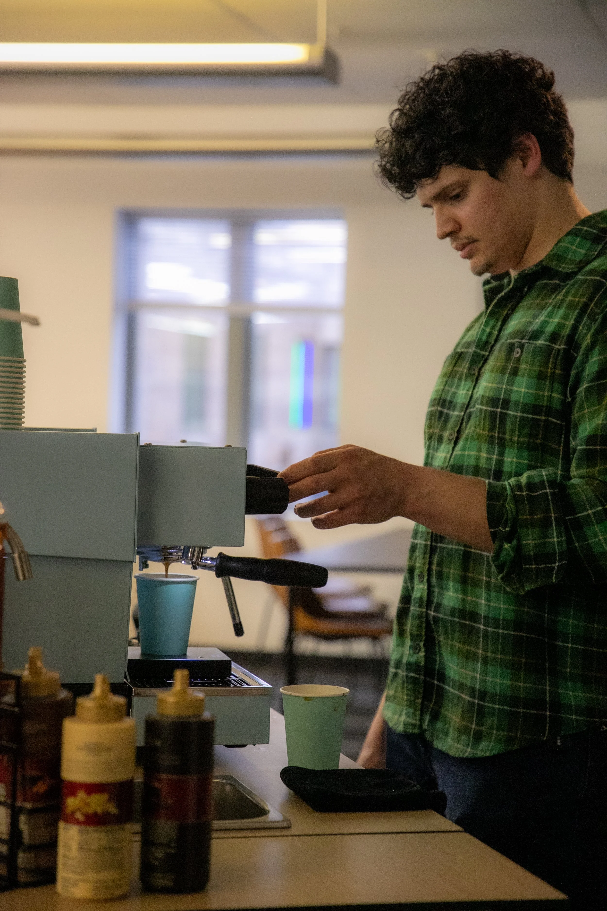 A man in a green checkered shirt making coffee with an espresso machine in a cafe or kitchen setting.