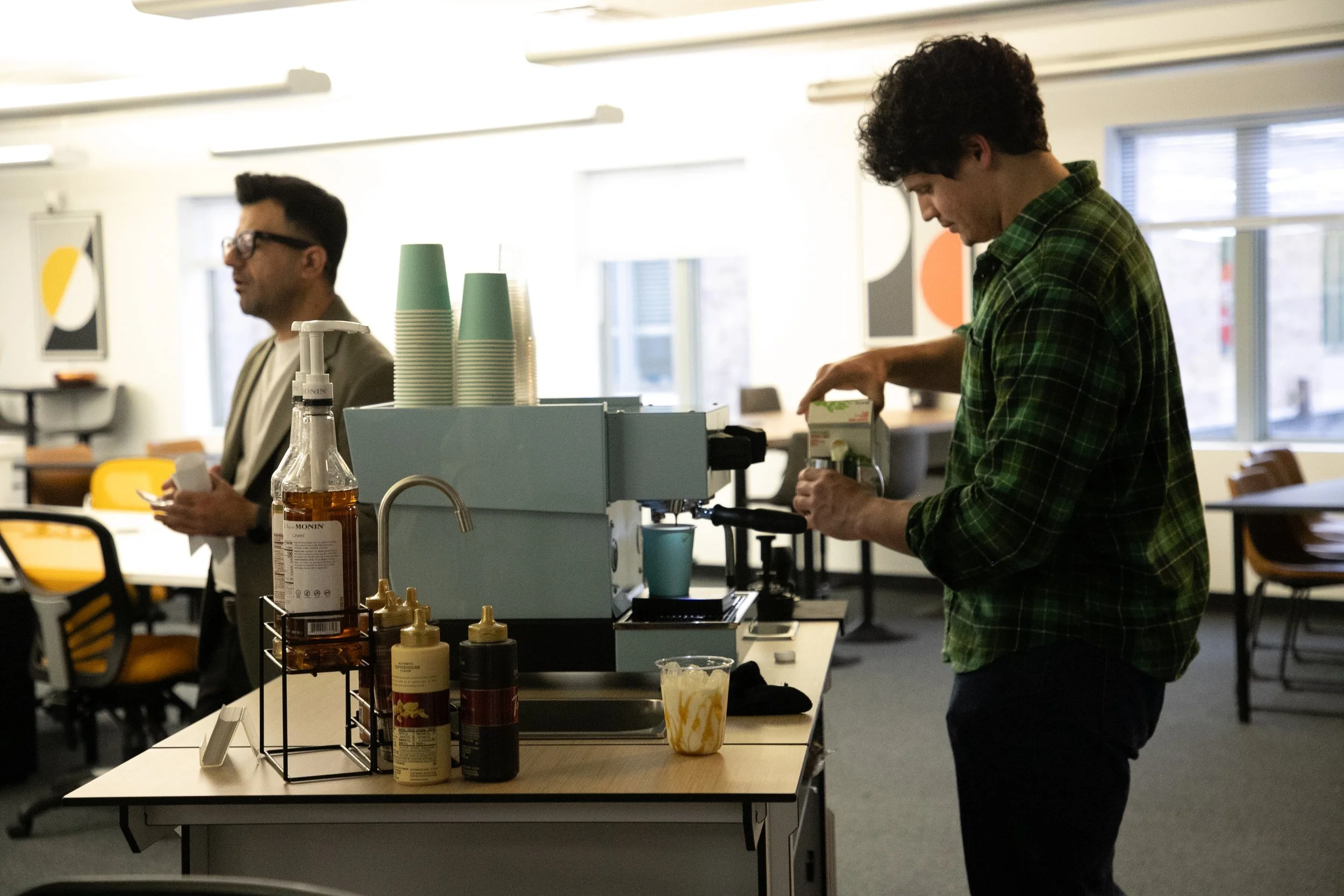 A person with curly hair wearing a green plaid shirt making coffee behind a counter, with another person wearing glasses and a beige blazer in the background in a modern coffee shop or cafe.