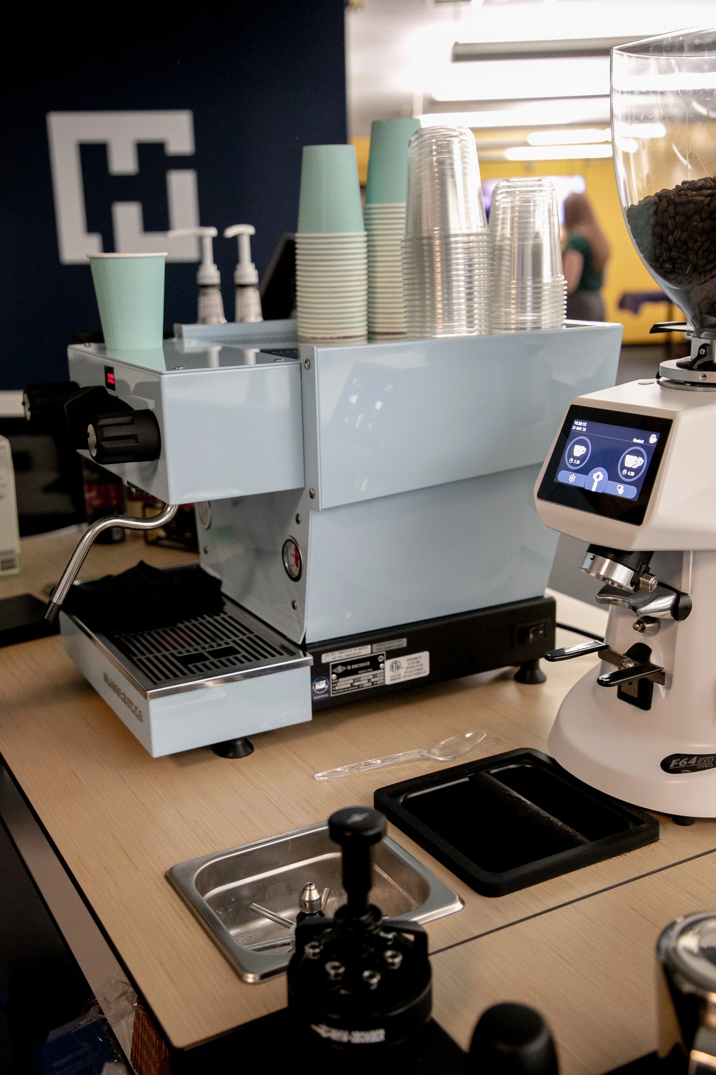 A close-up of a modern espresso machine on a wooden table in a coffee shop. There are paper cups stacked on top, a grinder to the right, and a touchscreen panel showing drink options. A sink is visible in the foreground.