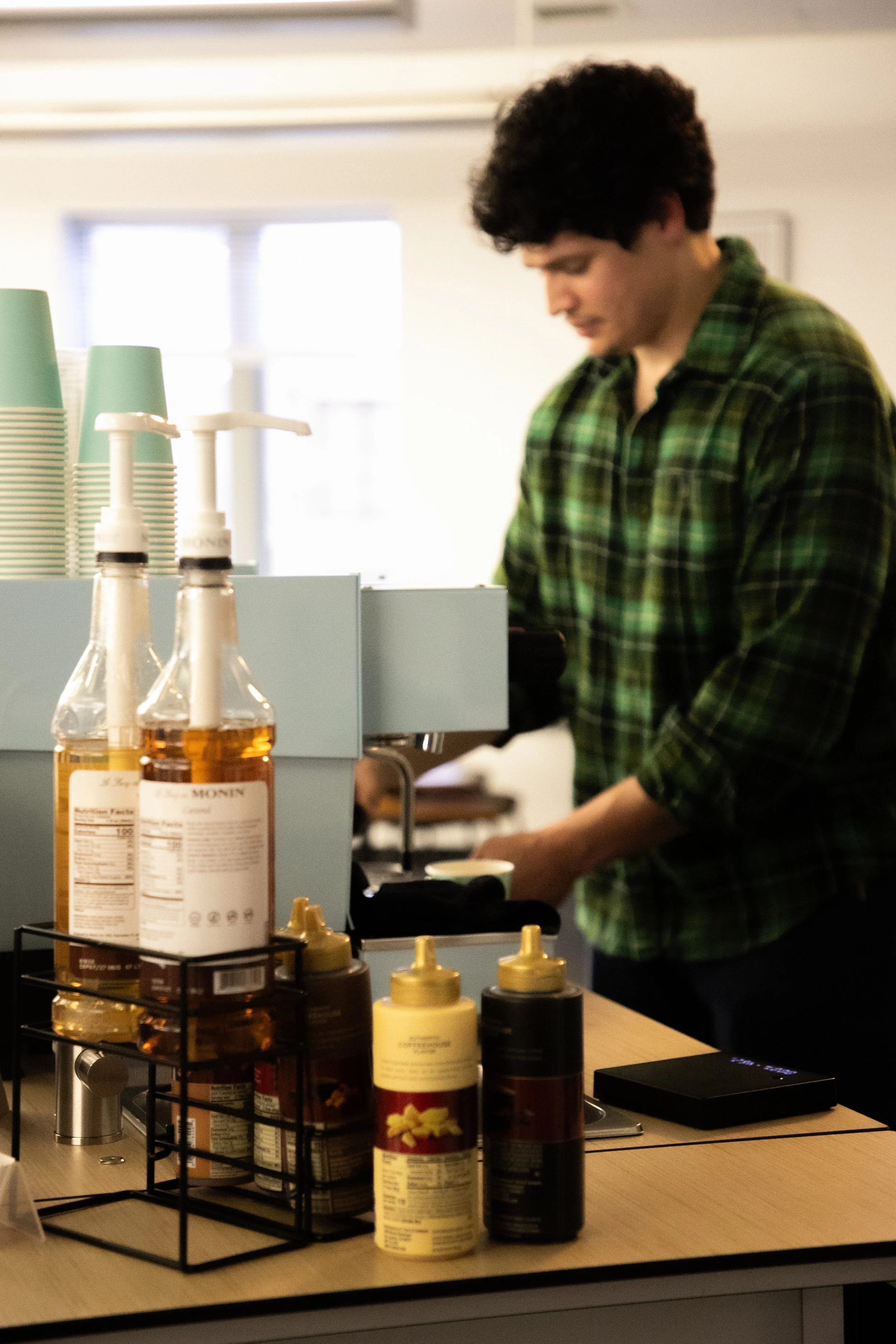 Young man with curly dark hair wearing a green plaid shirt, standing behind a counter in a café or coffee shop, looking down. In the foreground, there are syrup bottles and other condiments on a black wire rack on a light wooden counter.