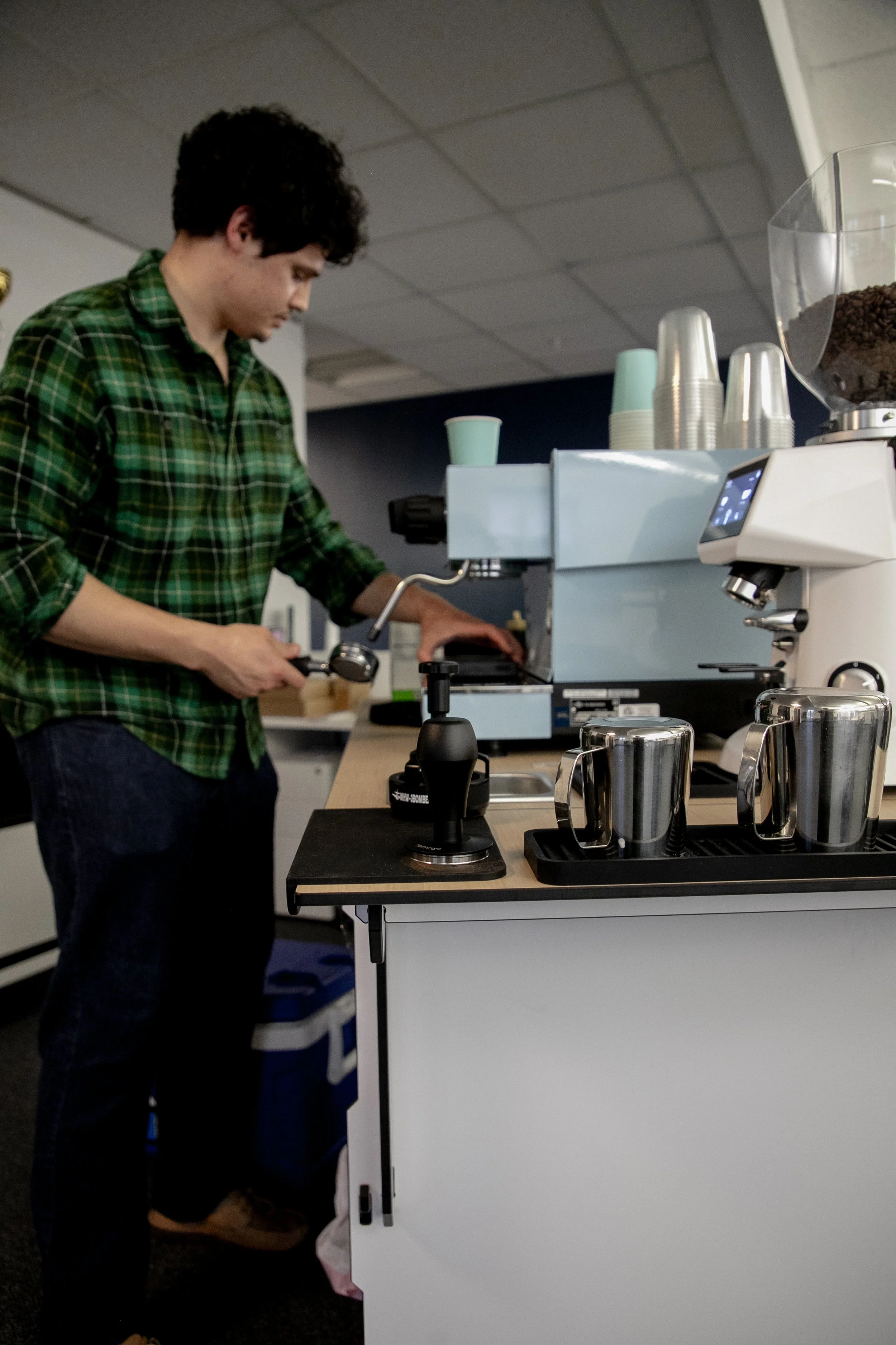 A person in a green plaid shirt preparing coffee in a commercial setting with a coffee machine, cups, and coffee beans on display.