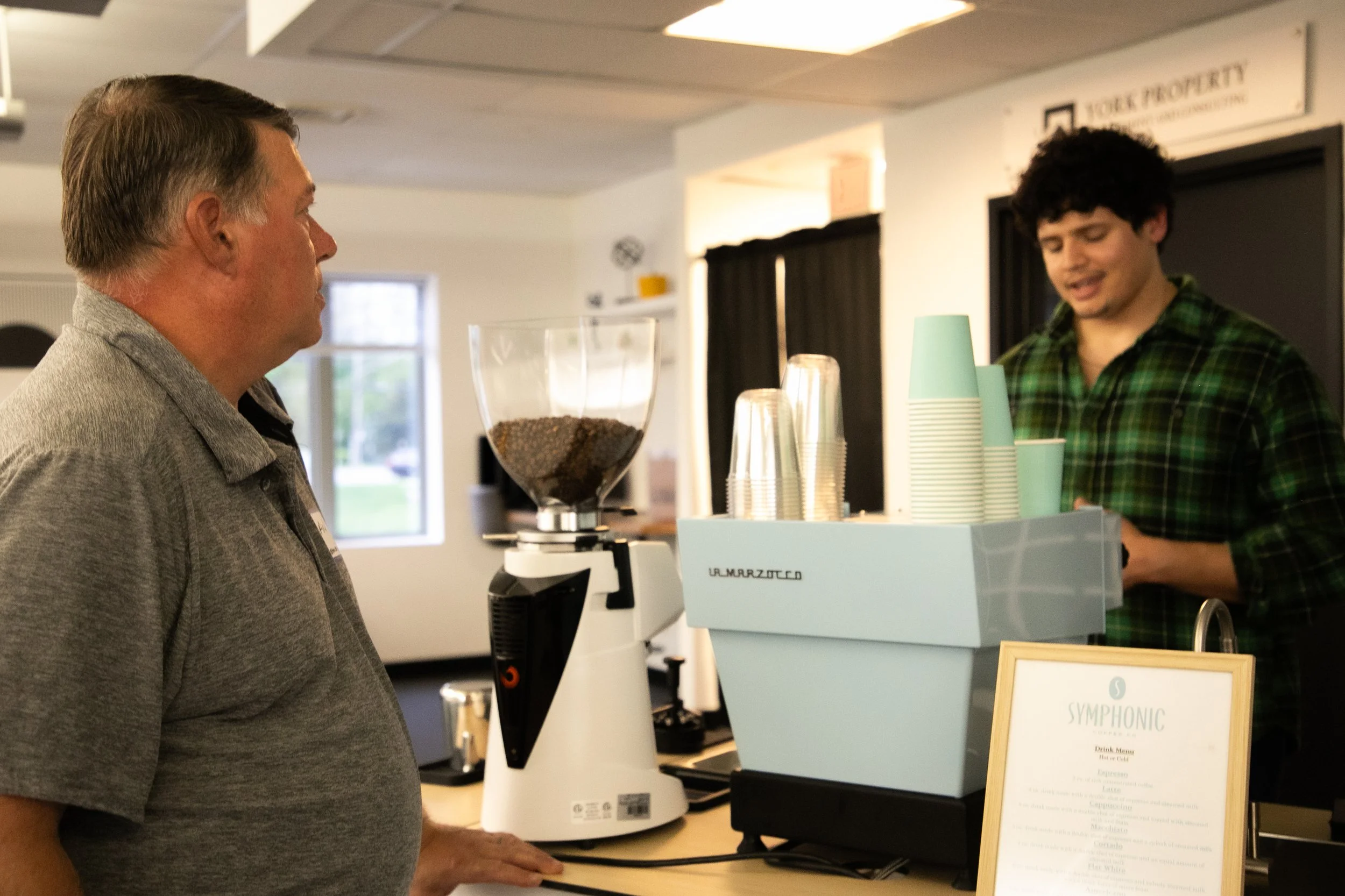 A man ordering coffee at a cafe counter with a barista behind the espresso machine, and a sign with the cafe menu on the table.