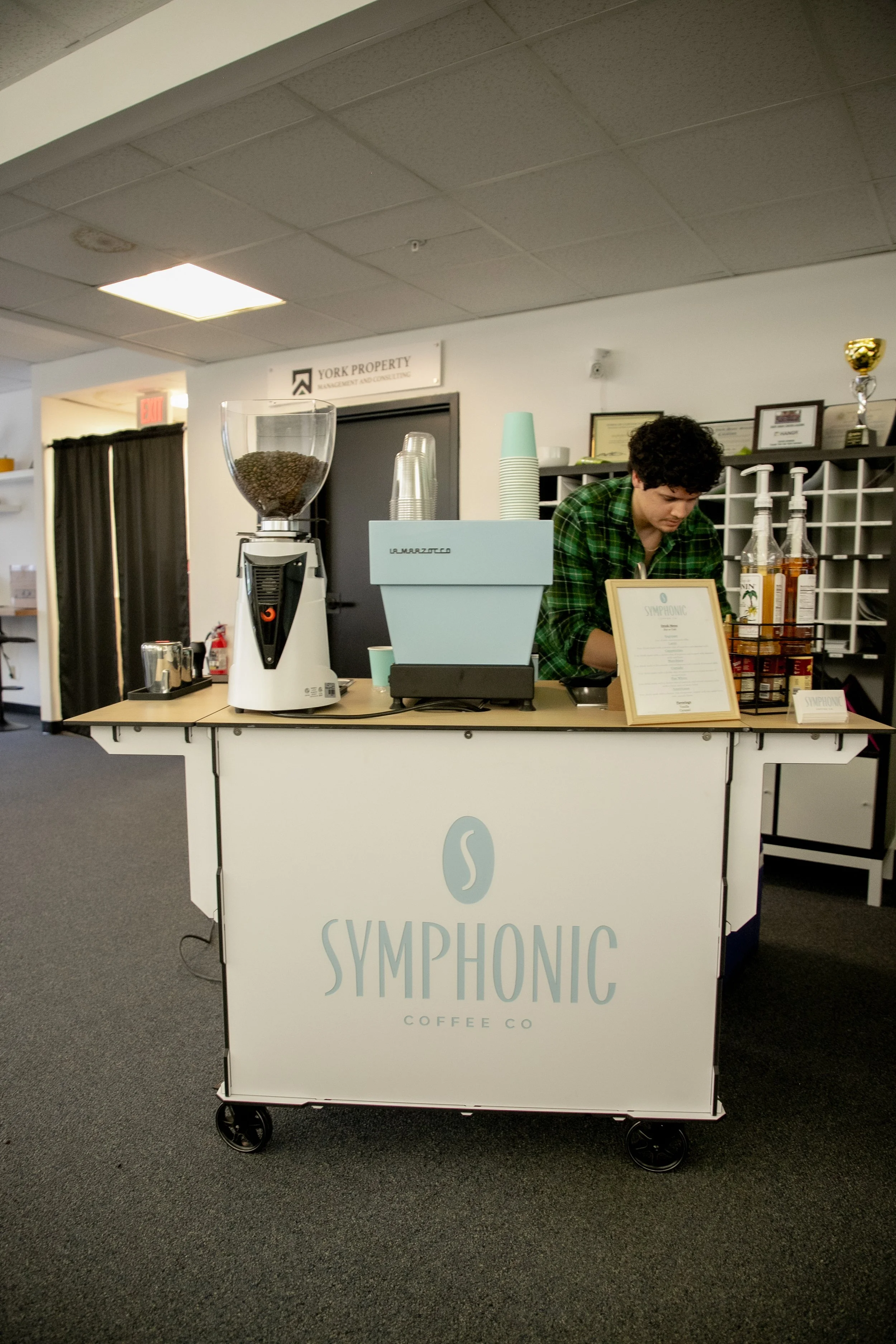 A coffee cart from Syphonic Coffee Co featuring a large sign, coffee beans in a grinder, bottles of syrups, and a barista with curly hair preparing a drink in an indoor setting with awards and certificates on the wall in the background.