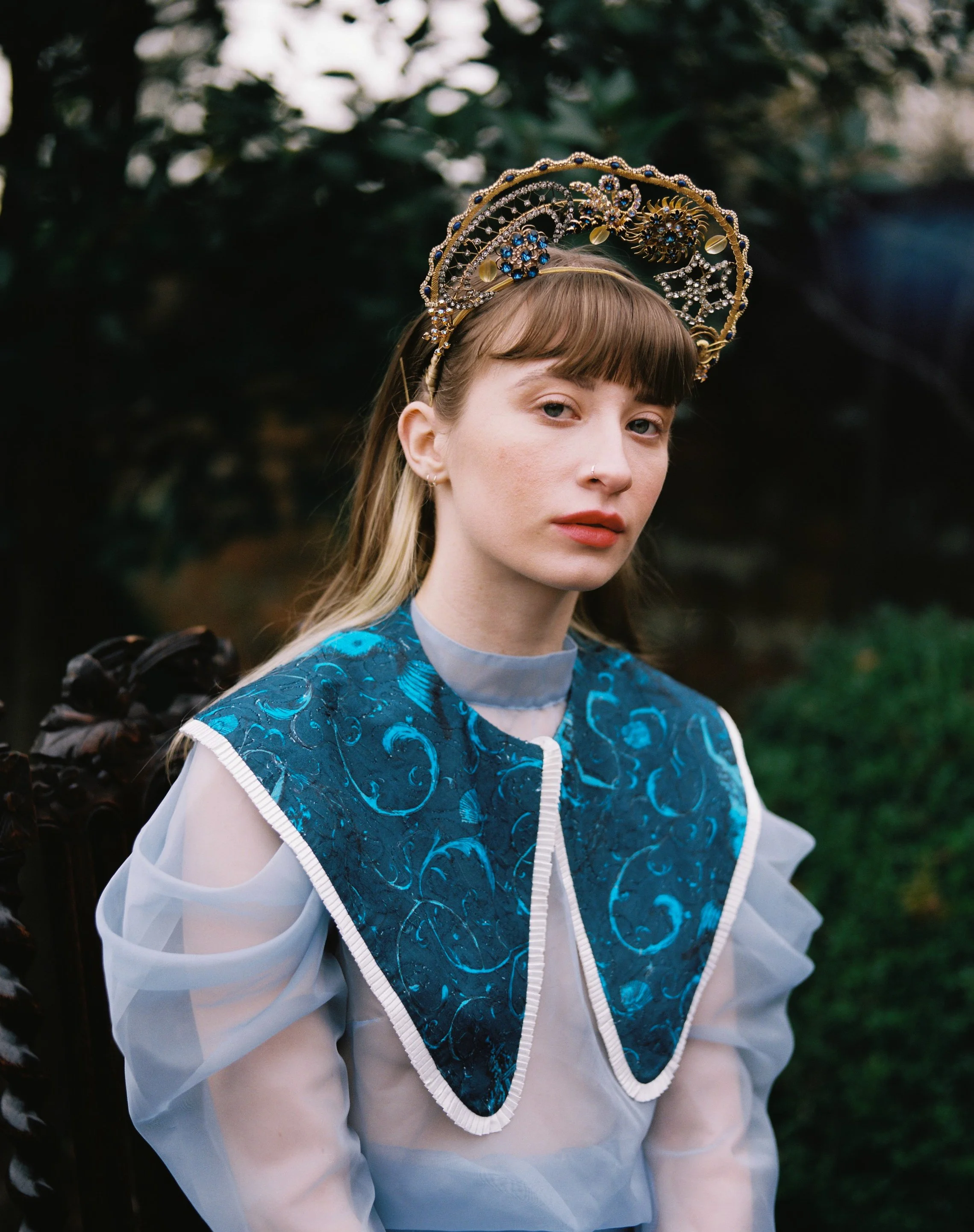 A young woman with light brown hair and bangs, wearing a blue embroidered collar and sheer white sleeves, seated outdoors with greenery in the background. She has a decorative gold and jewel crown on her head.