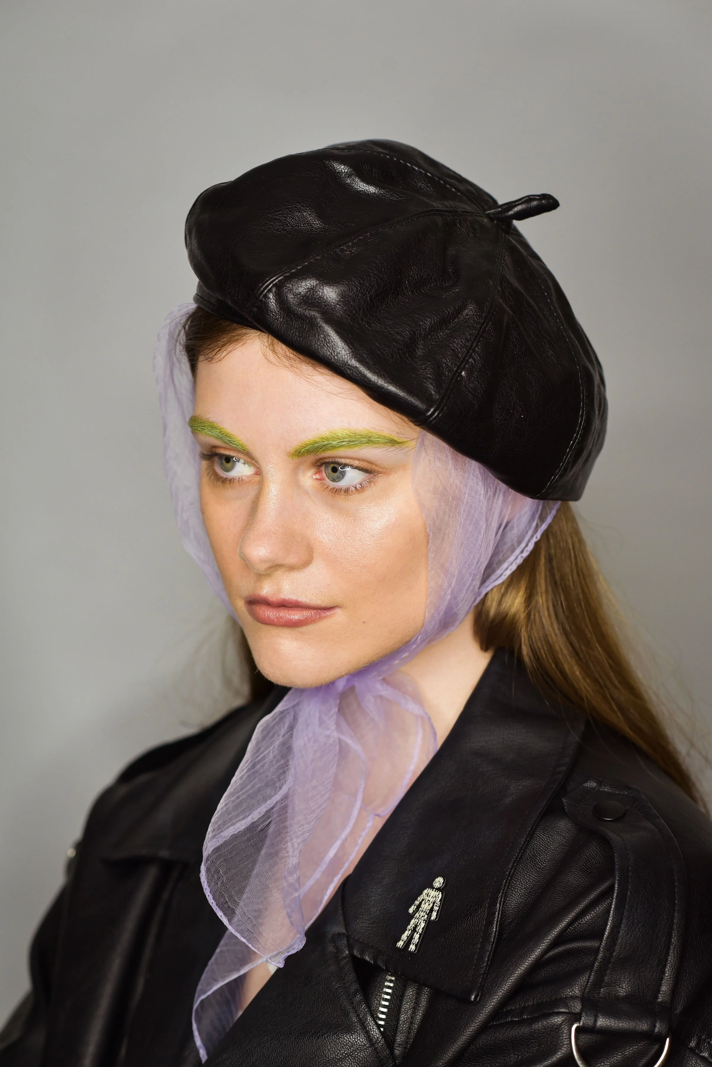 A woman with green eyebrows, wearing a black leather beret, purple sheer scarf, and black leather jacket with miniature figure embroidery.