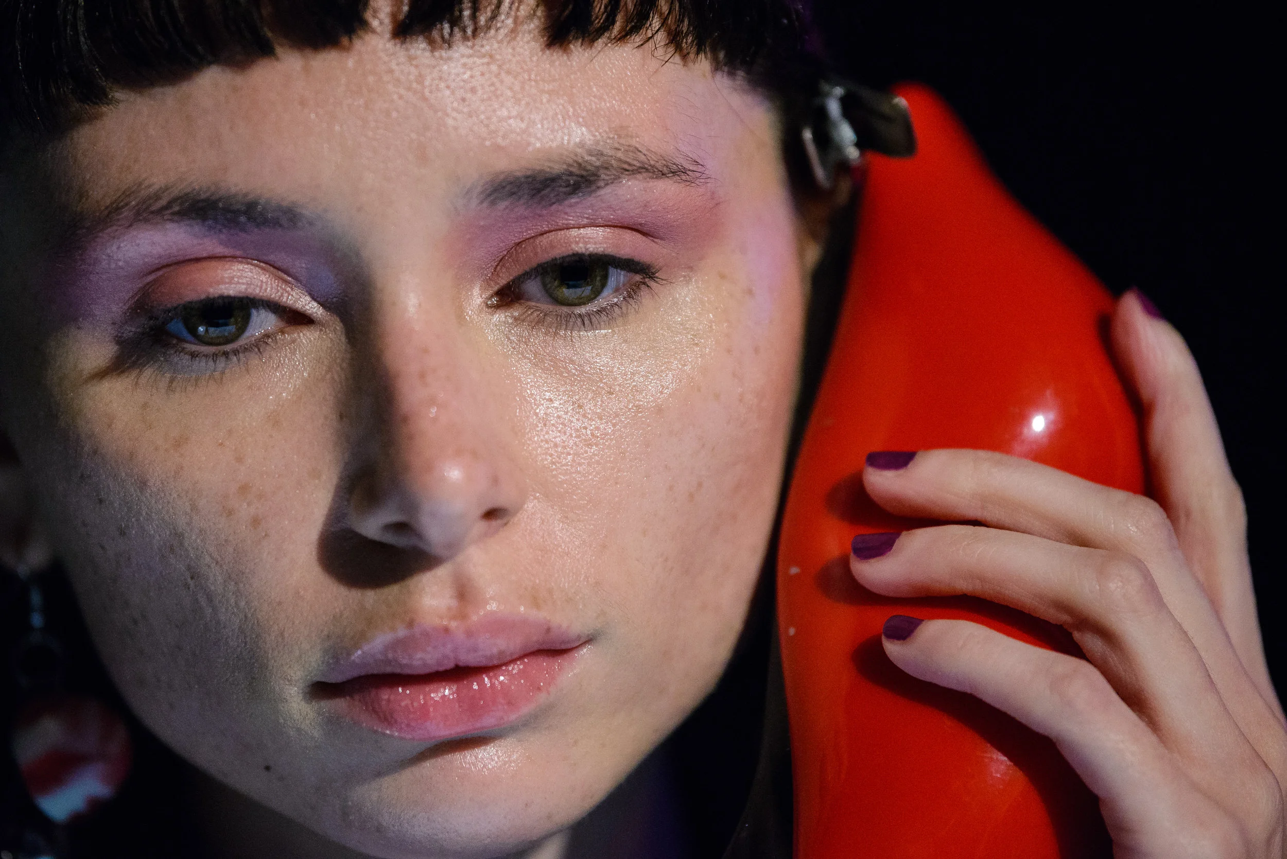 Close-up of a woman with makeup, holding a red vintage telephone to her ear, with a black background.