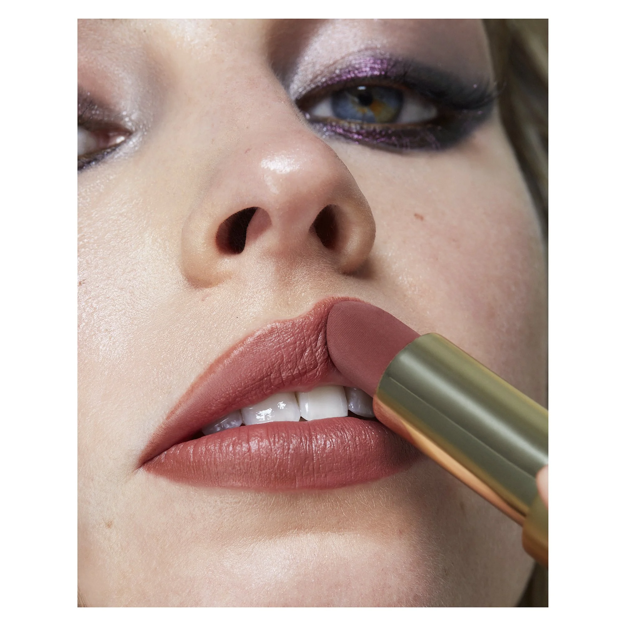 Close-up of a woman's face applying nude lipstick with a gold-tipped lipstick tube, makeup including pink and purple eyeshadow visible.