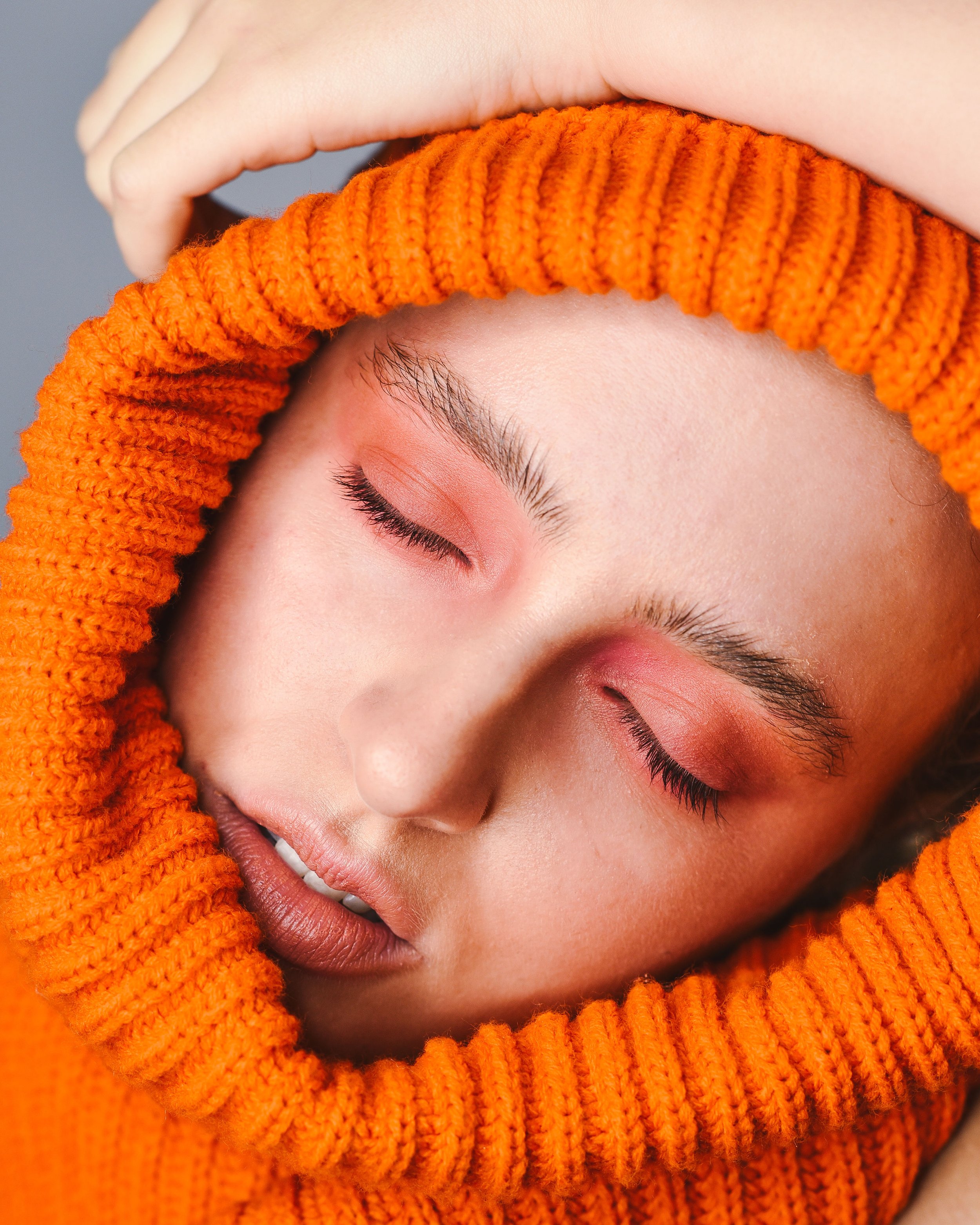 A close-up of a woman with closed eyes, wearing an orange knitted hood or sweater, with makeup in warm tones.