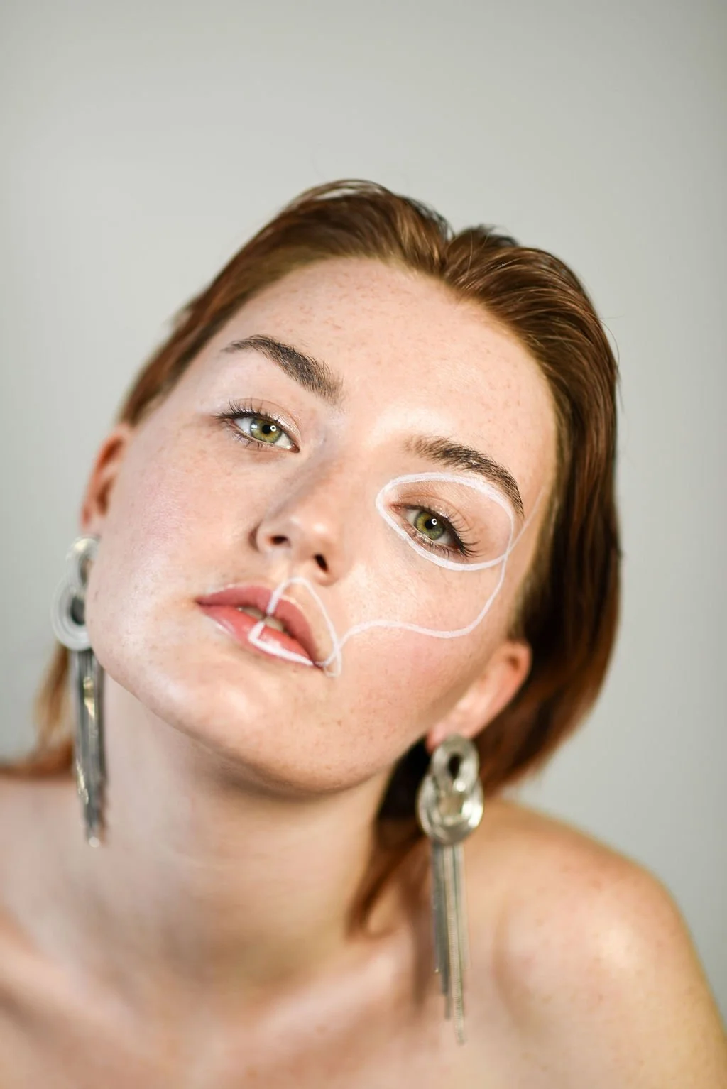 A woman with red hair and green eyes looking at the camera with white lines drawn on her face, indicating makeup or cosmetic planning.