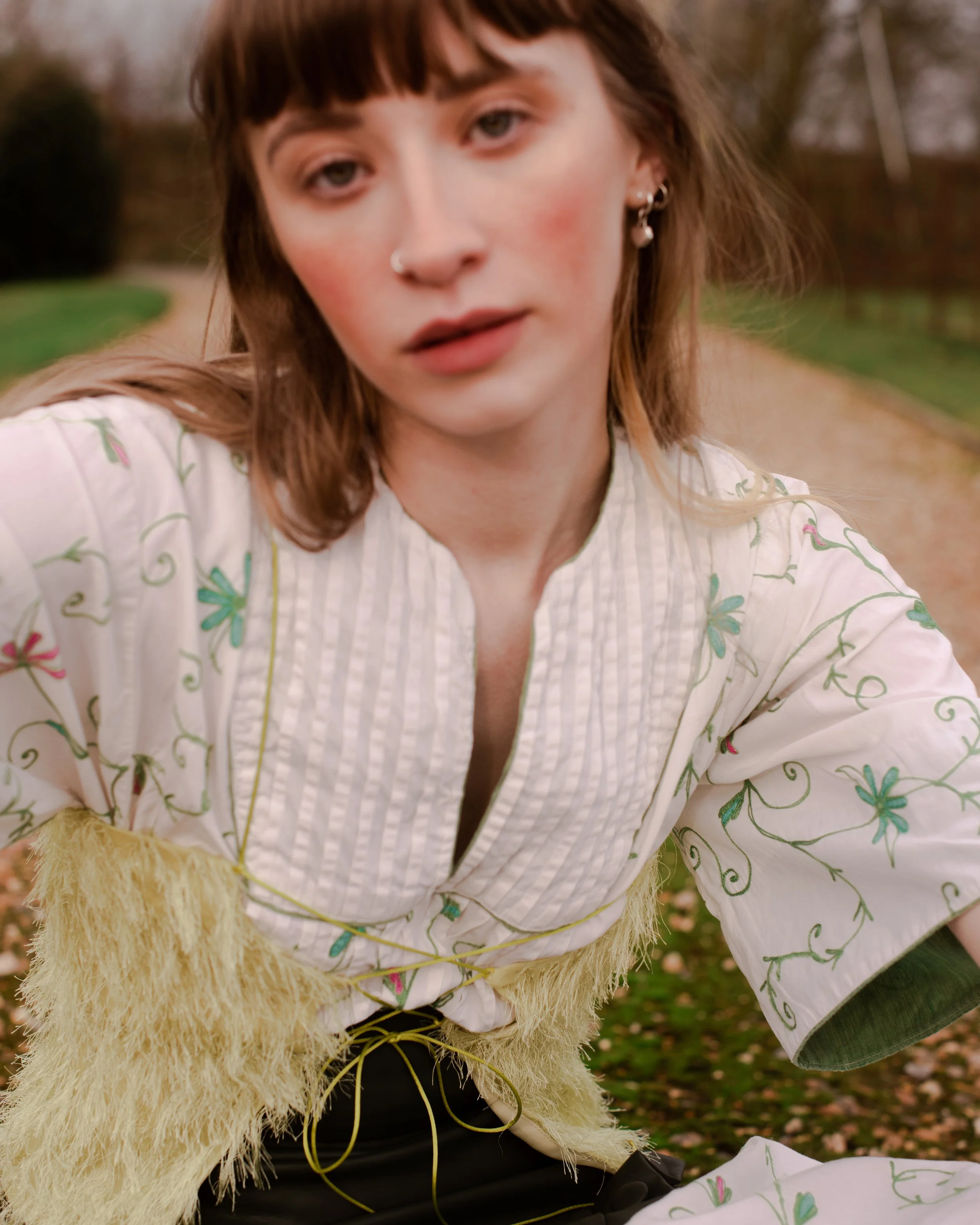 Close-up of a young woman with light skin, shoulder-length brown hair, and hoop earrings, wearing a white embroidered top and a yellow fuzzy vest, outdoors on a pathway in a park or garden.