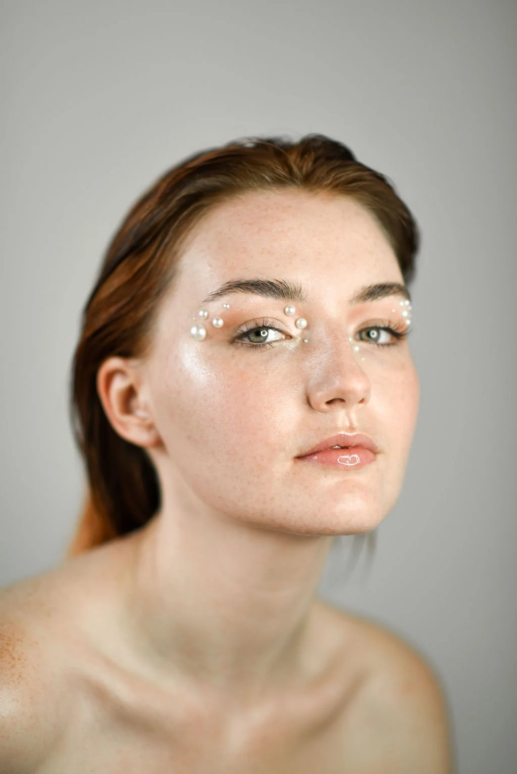 A woman with auburn hair and fair skin with freckles, looking at the camera. She has pearl embellishments around her eyes and a glossy lip, against a plain light gray background.