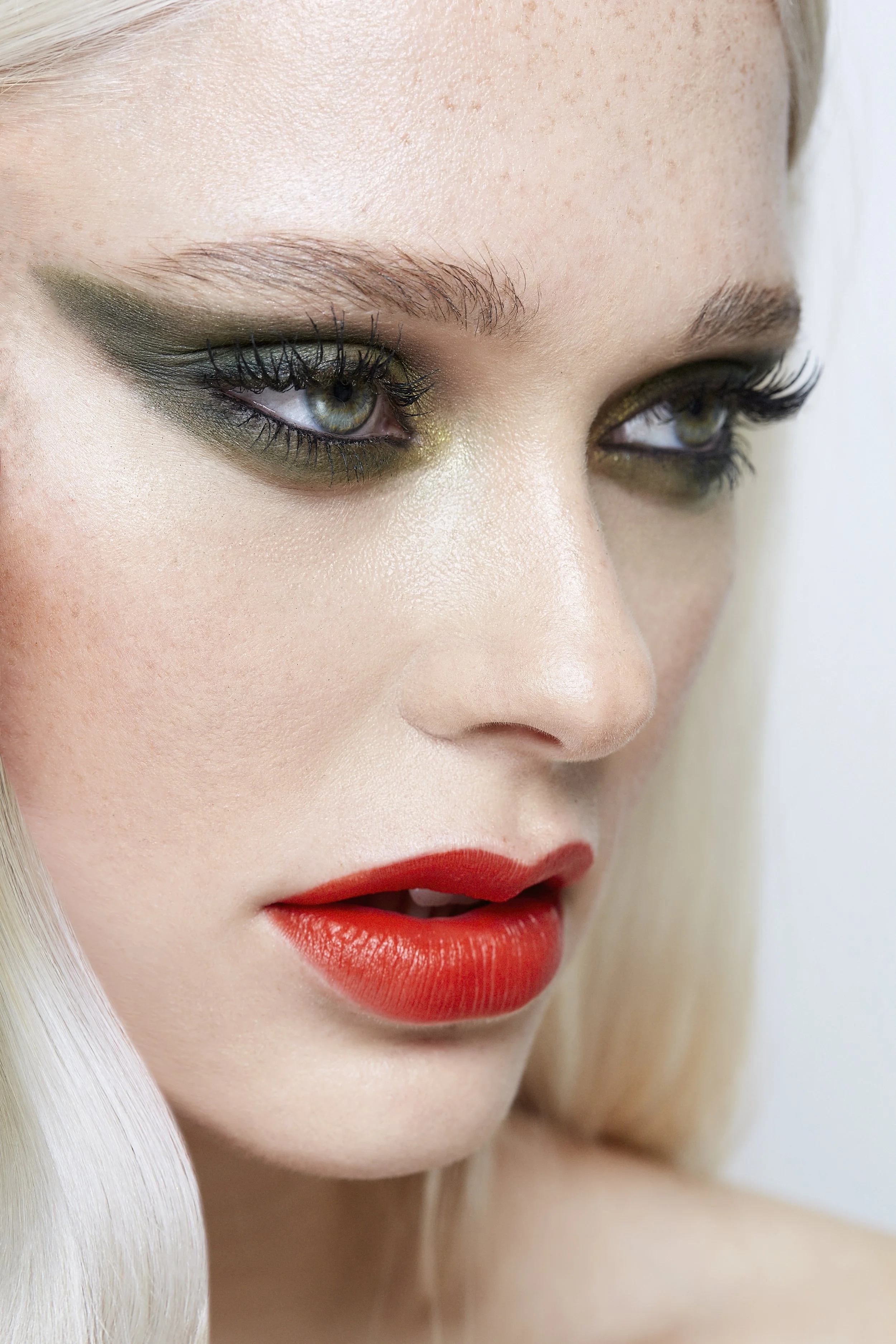 Close-up of a woman's face showcasing her eye makeup with dark green eyeshadow, long eyelashes, and colorful eyes, along with red lipstick and fair skin.