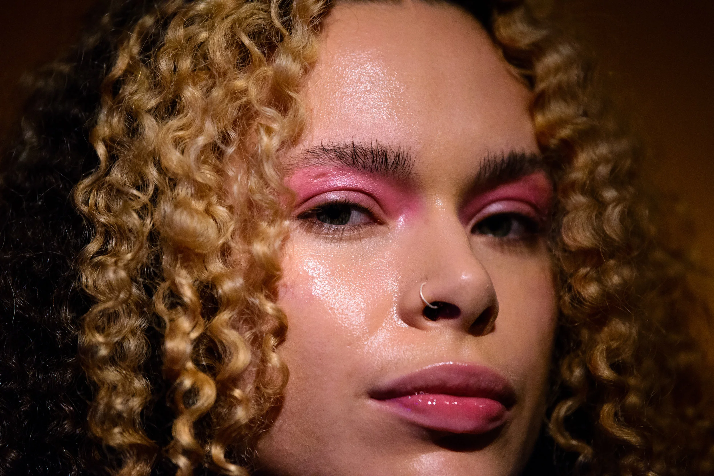 A close-up of a woman with curly blonde hair, wearing pink eyeshadow, glossy lips, and a nose ring.