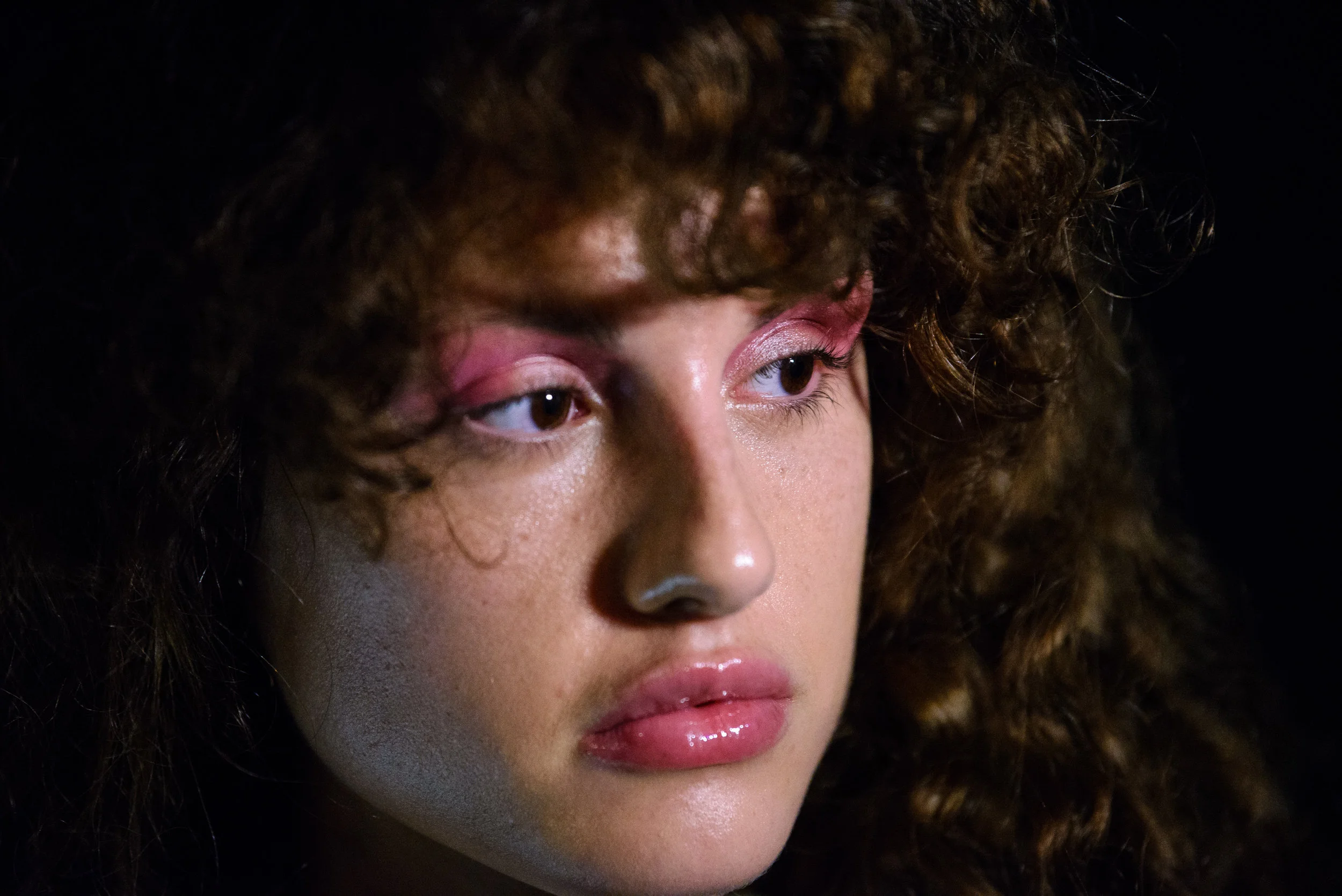 Close-up of a woman's face with curly hair and makeup featuring pink eyeshadow and glossy lips.