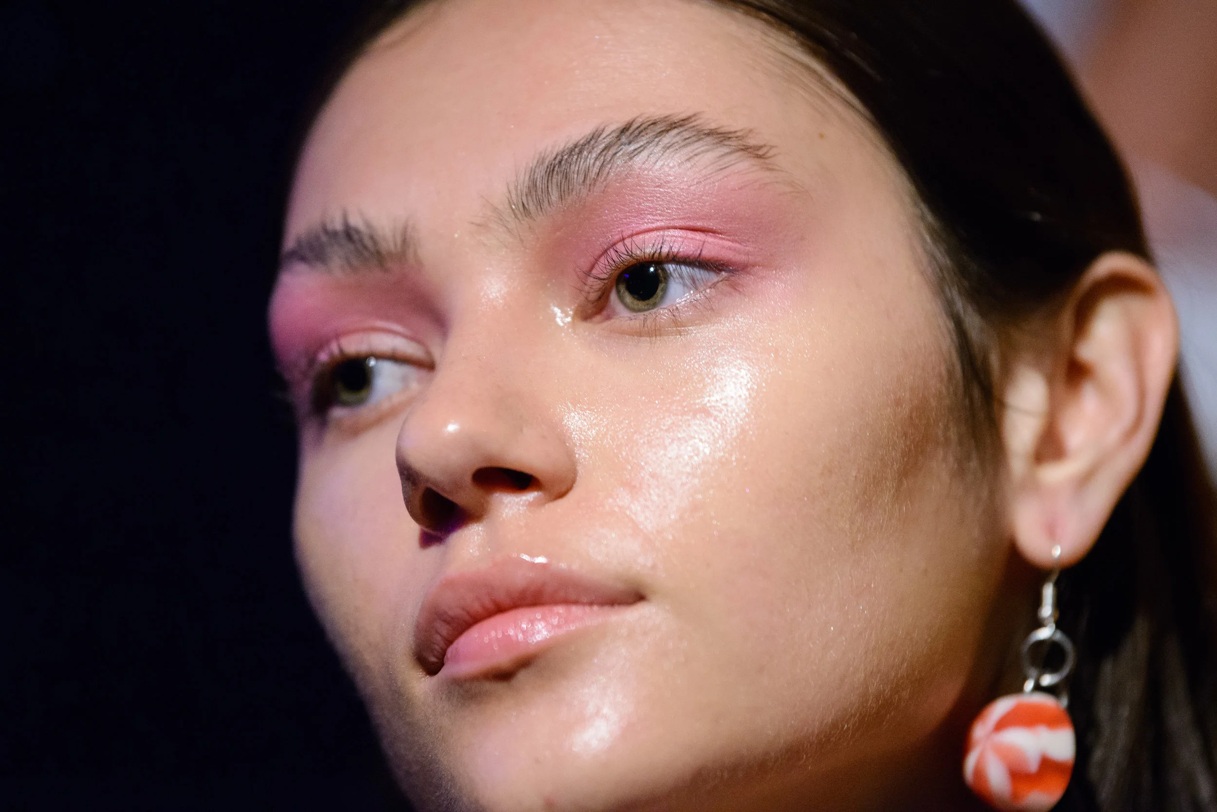 Close-up of a woman with pink eyeshadow, dewy skin, minimal makeup, and a black hairstyle, wearing a silver hoop earring with an orange and white flower pendant.