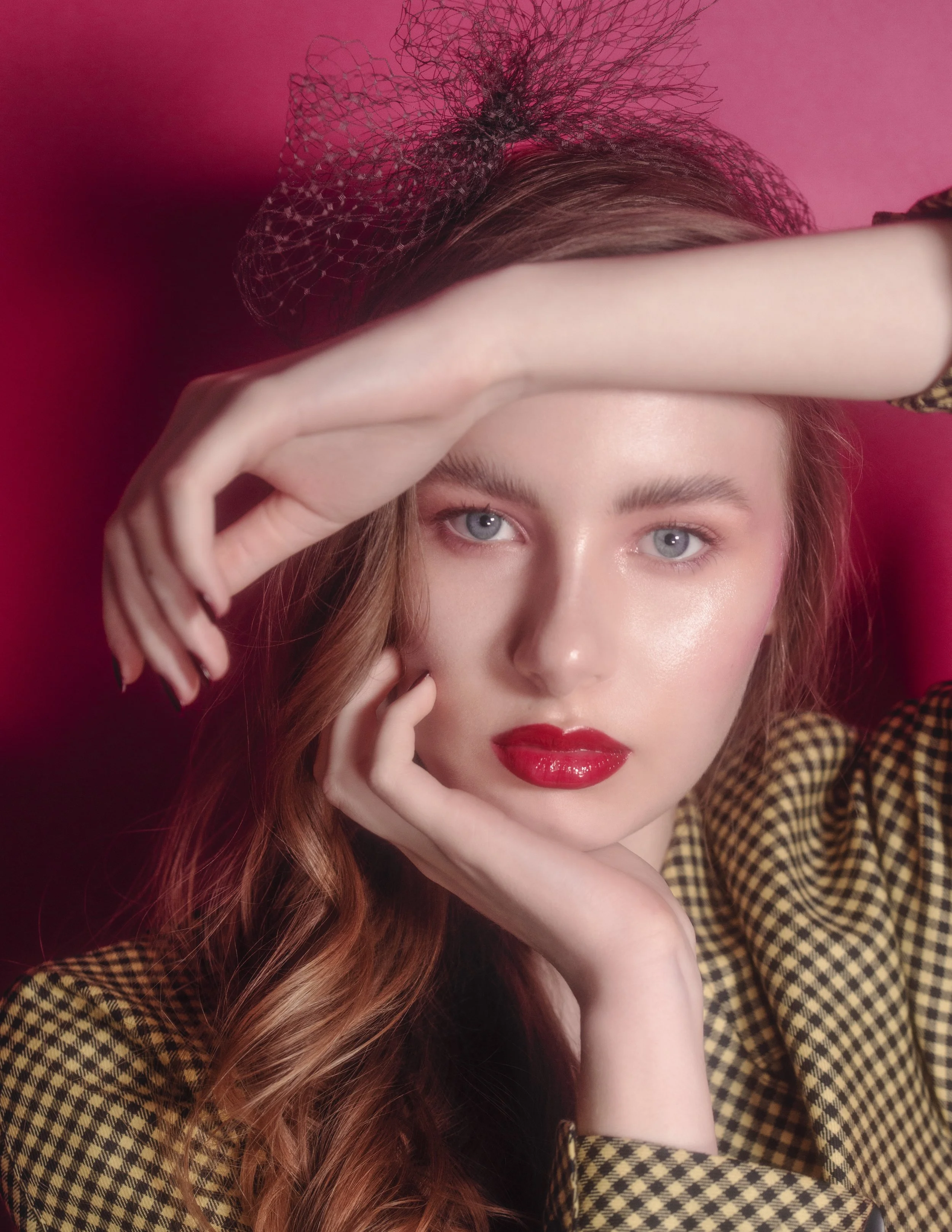 A young woman with red hair, wearing red lipstick and a black mesh hat, posing against a pink background, touching her forehead with one hand and her chin with the other.