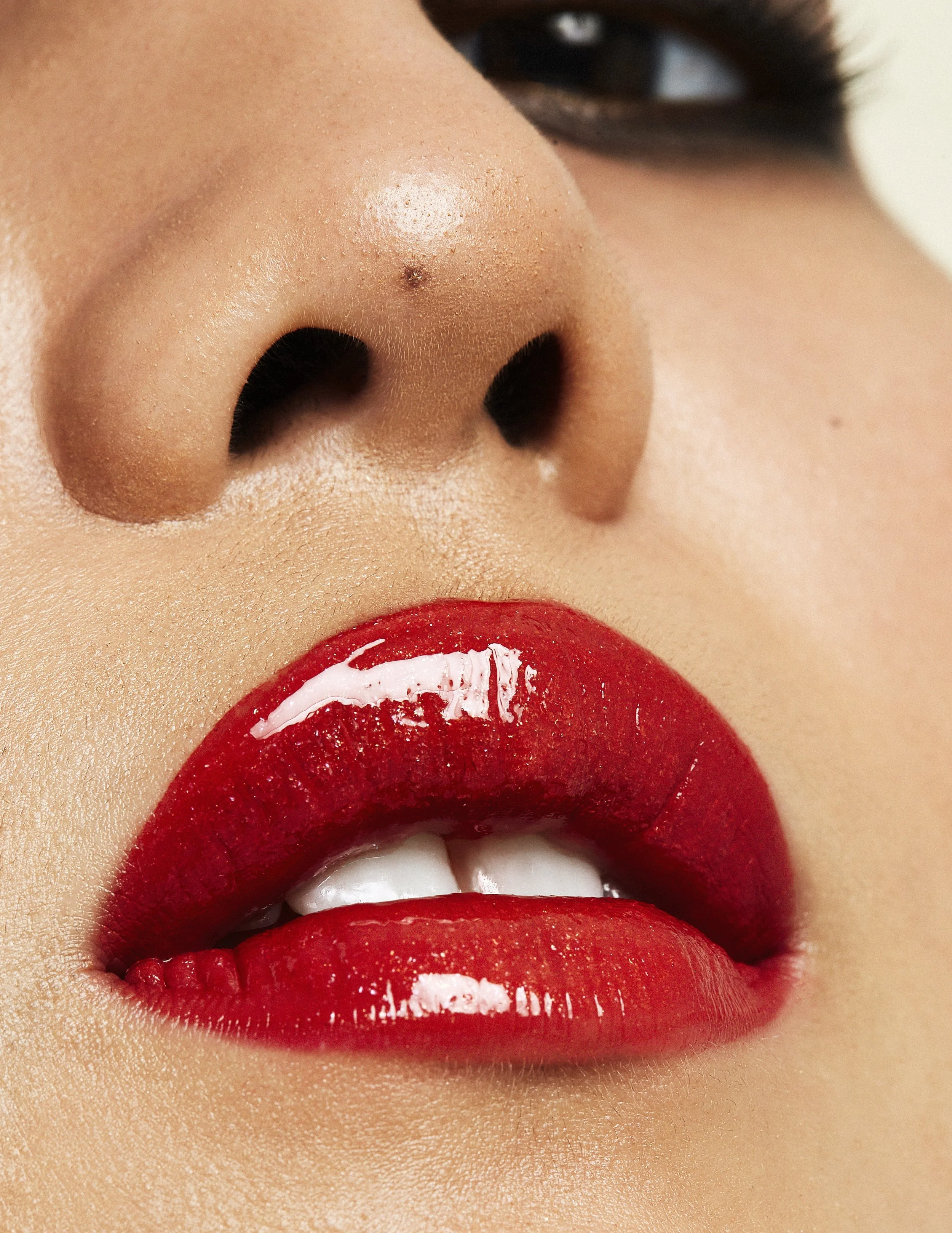 Close-up of a person's lips painted with shiny red lipstick, nude skin, and a visible nose with a beauty mark.